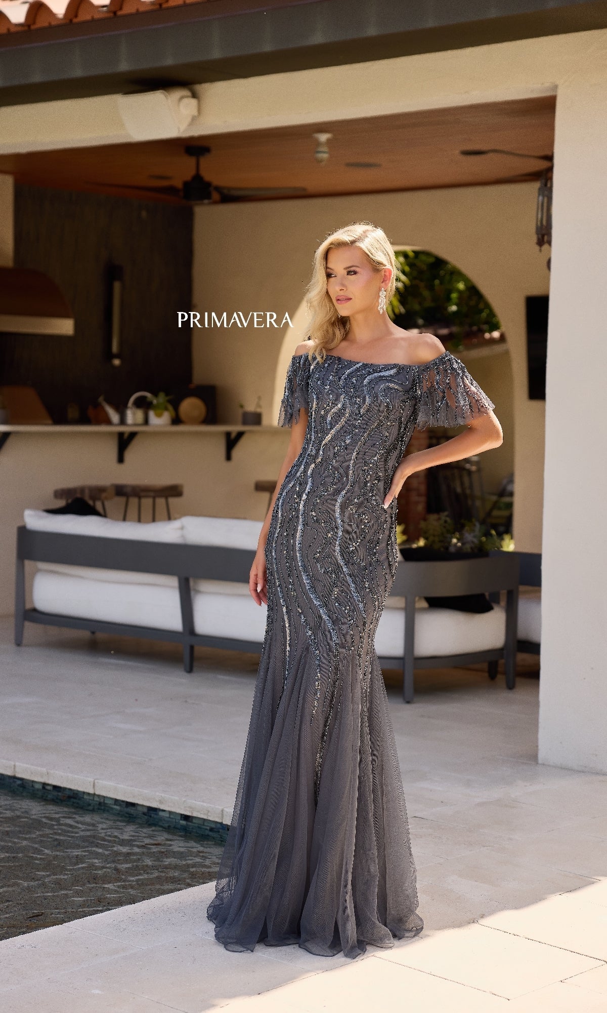 A woman stands poolside on a modern patio, wearing the Long Formal Dress: Primavera 13155—a fitted, off-the-shoulder sequin mermaid gown with sheer embellished sleeves that shimmers elegantly in the light.