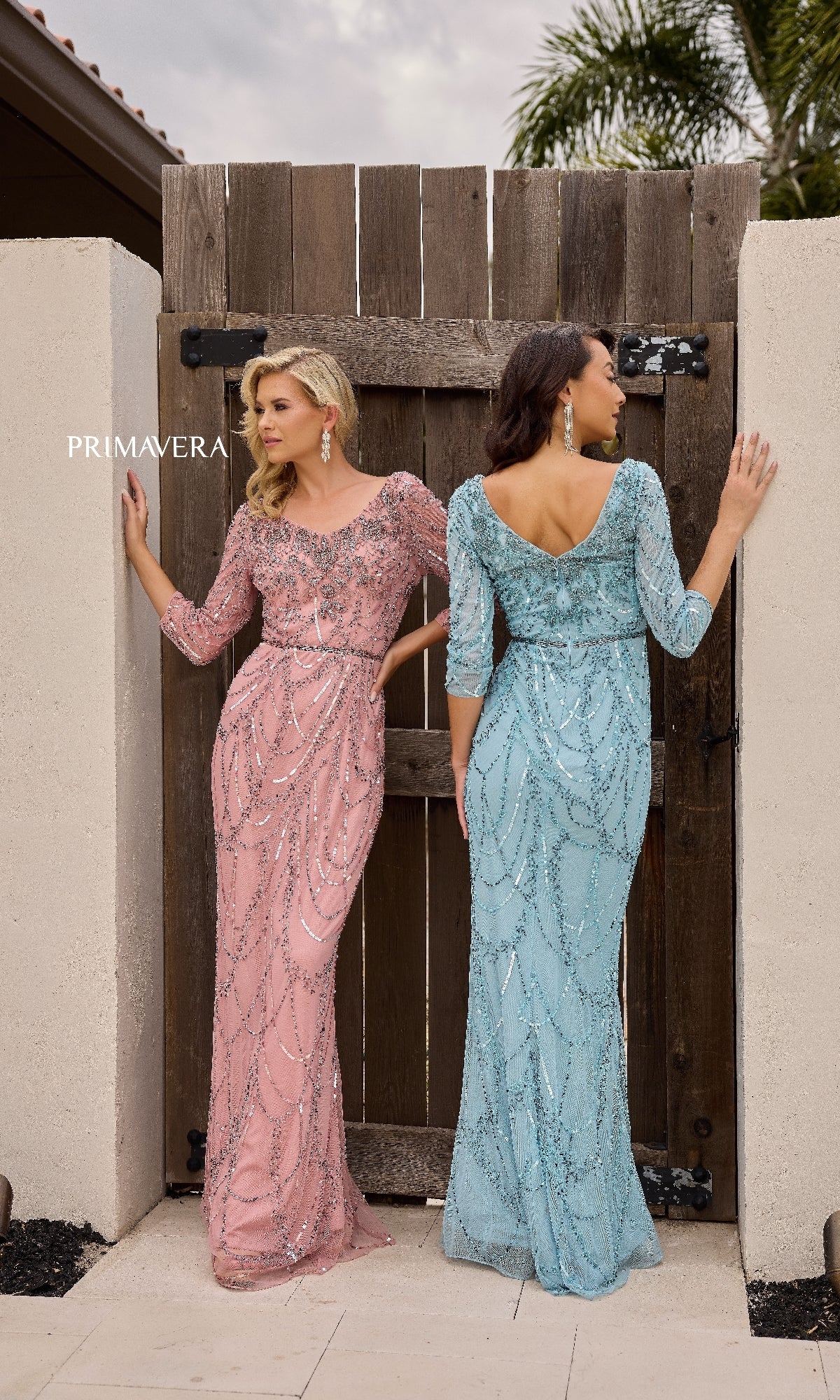 Two women stand by a wooden gate wearing Primavera 13156 long formal dresses—one in pink, facing forward, and one in light blue with a beaded waistline, facing away.
