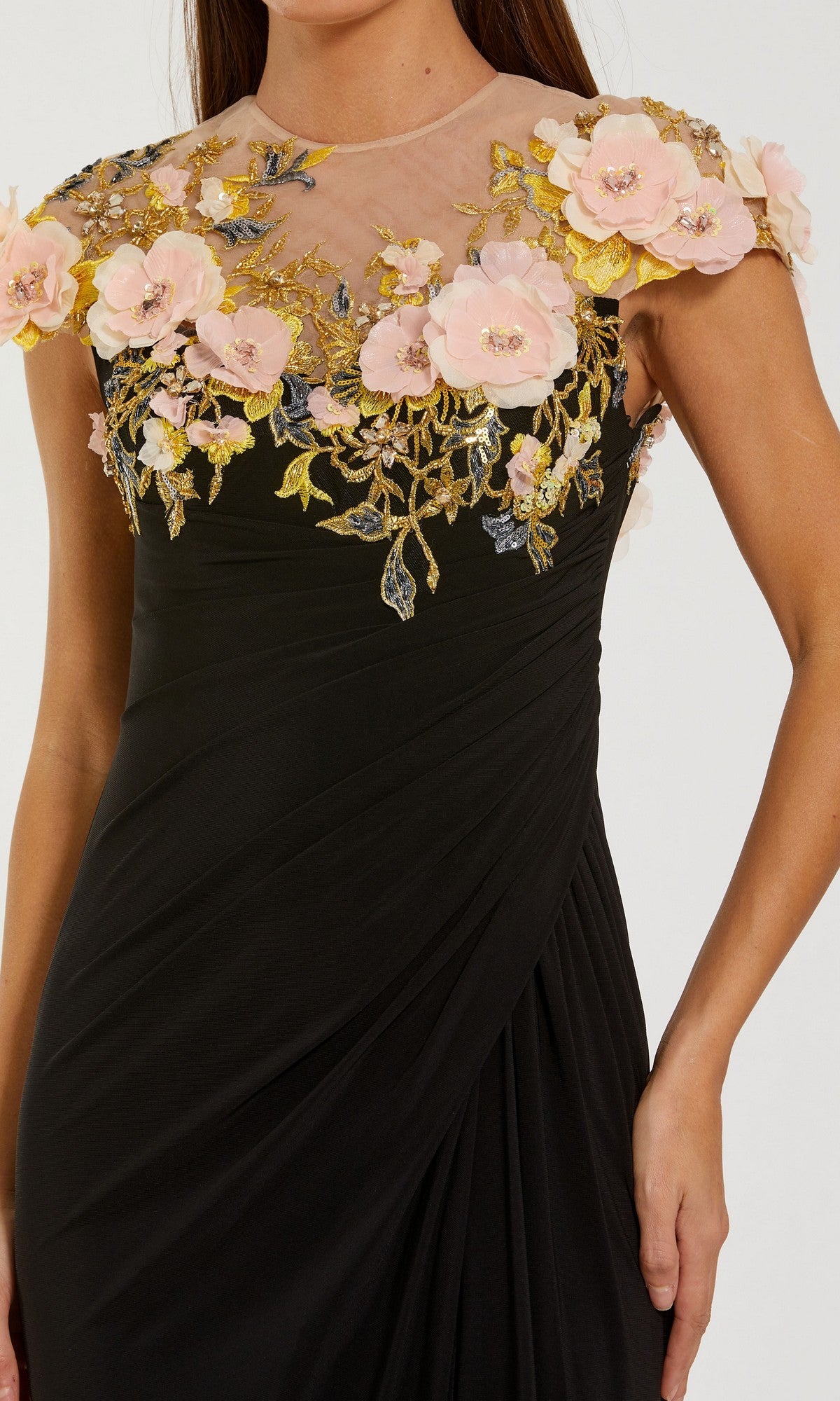 A woman is wearing the Mac Duggal 13207 long formal dress, featuring a black sheer neckline, pink 3D floral accents, gold embroidery, and cap sleeves. Only her upper body is visible.