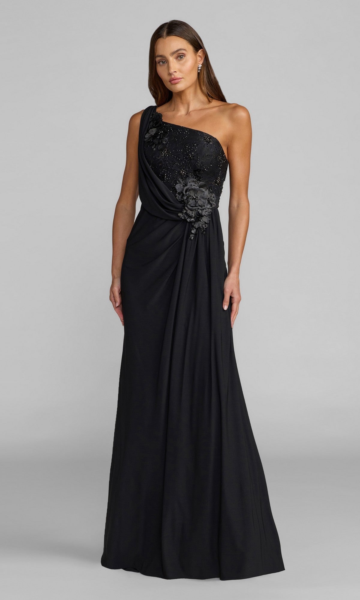 A woman wearing the Mac Duggal 13210 long formal dress in black, featuring a one-shoulder design with 3D flower detailing and elegant draping, stands against a plain gray background.