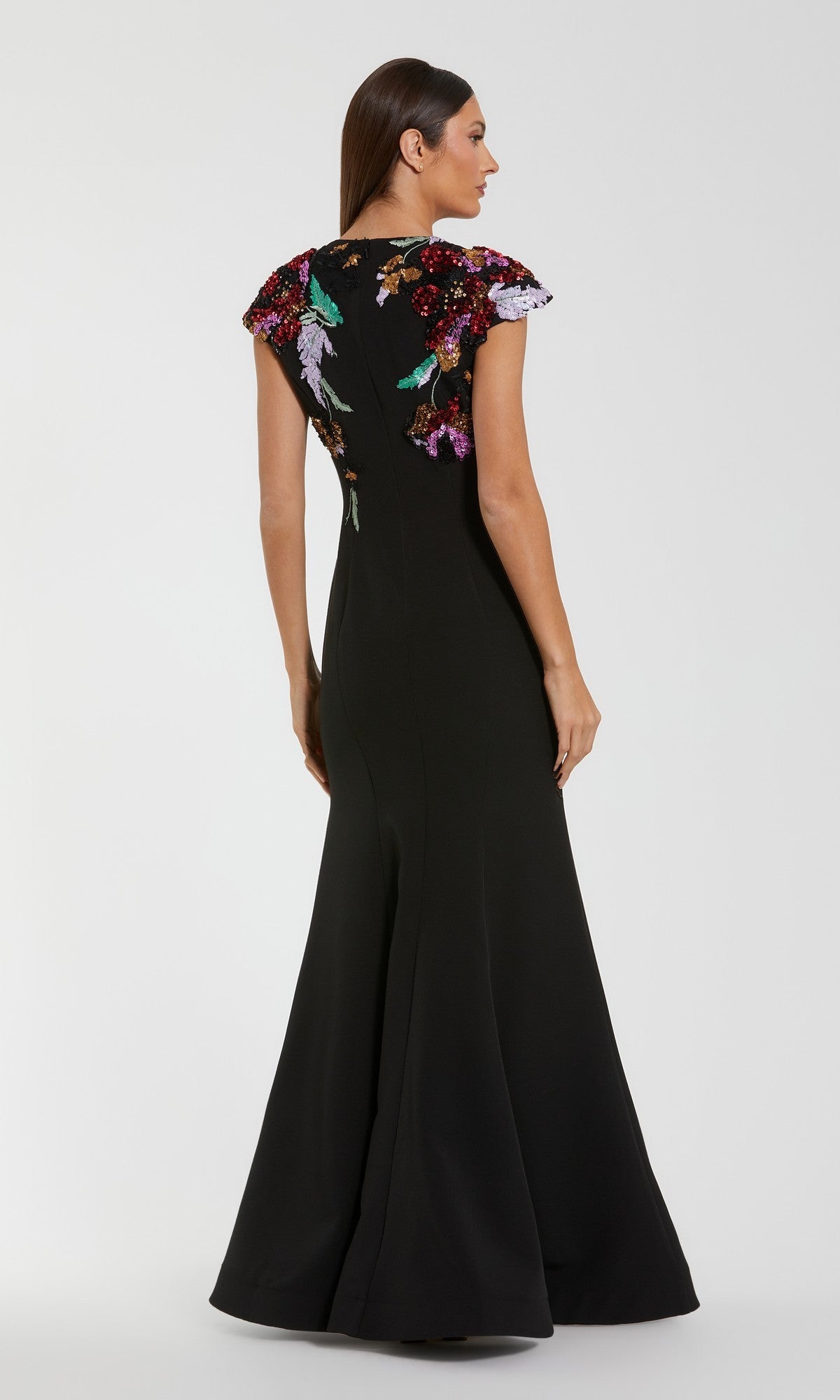 A woman stands facing away from the camera, wearing the Mac Duggal 13211 long formal dress in black crepe, featuring colorful floral sequin embroidery on the shoulders and upper back.