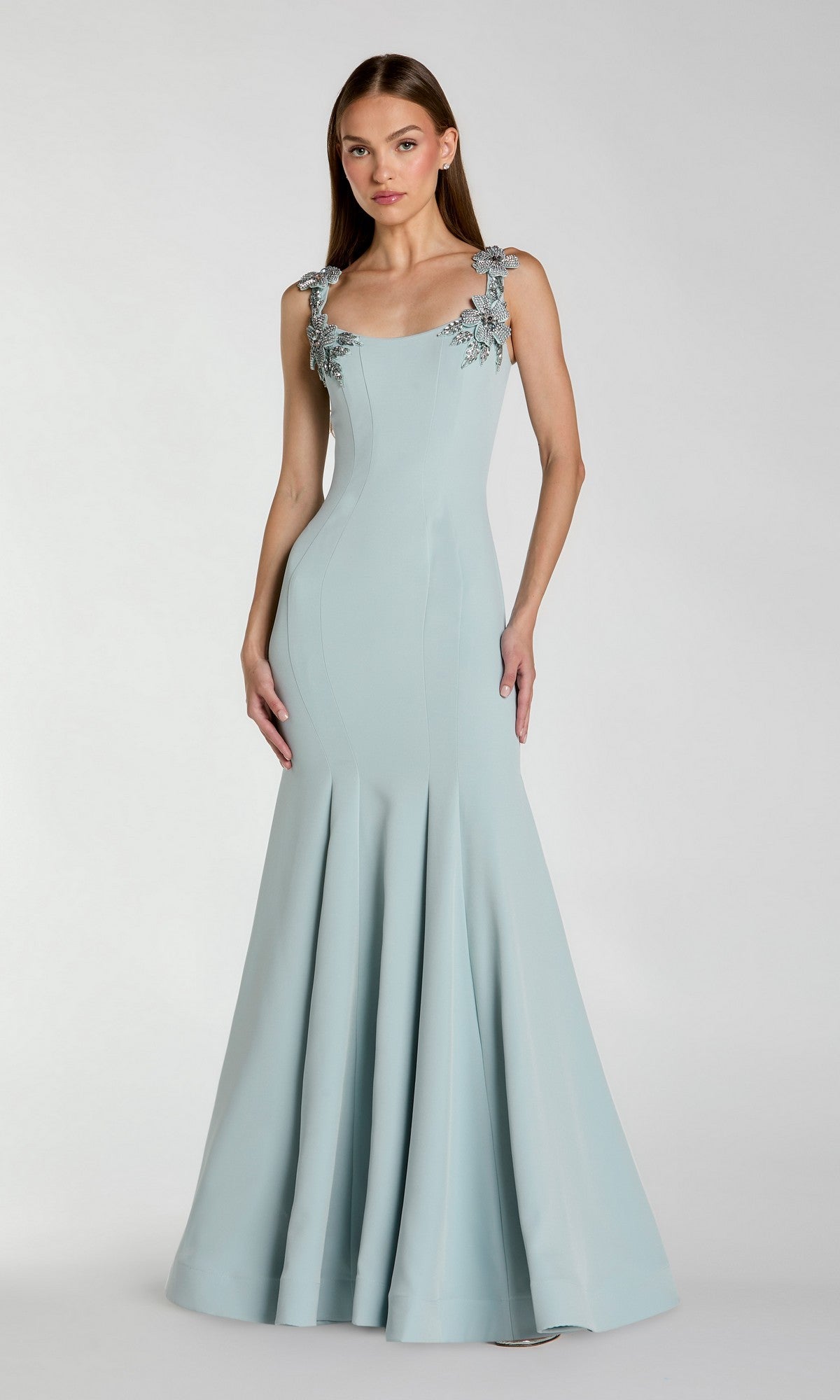 A woman stands against a plain light background, wearing the Mac Duggal 13224 Long Formal Dress in powder blue—a floor-length gown featuring floral embellishments on the straps.