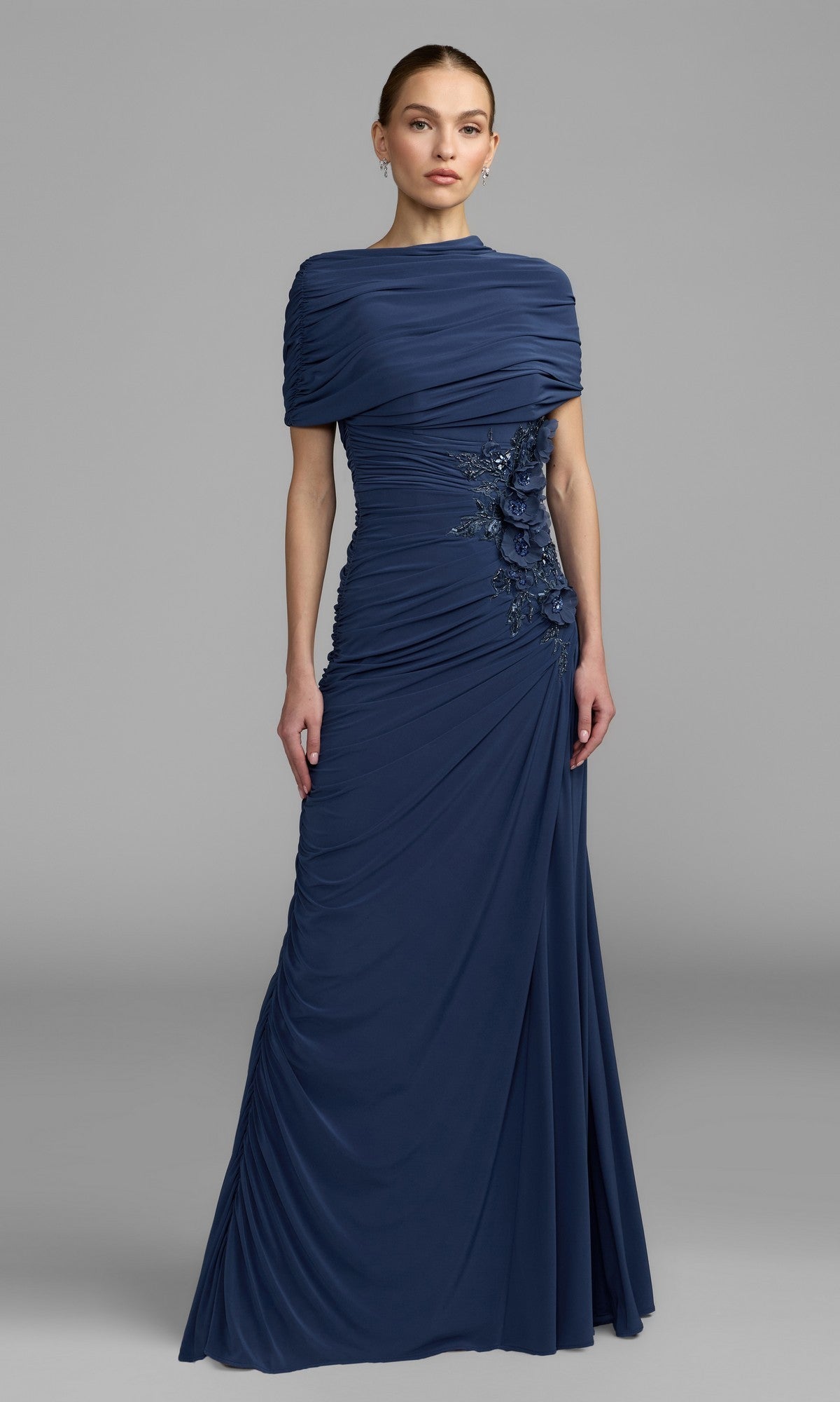 Dressed in the Mac Duggal 13244 long formal dress in stunning sapphire, a woman stands against a plain gray background. The floor-length gown features side embellishments and elegant ruched detailing—making it a sophisticated choice for a mother-of-the-bride.