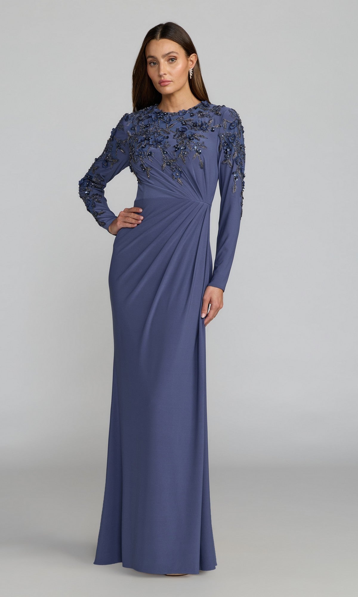 A woman wearing the Mac Duggal 13246 long formal dress in Blue Haze, featuring floral embroidery on the bodice and sleeves and styled with elegant modesty, stands against a plain light gray background.