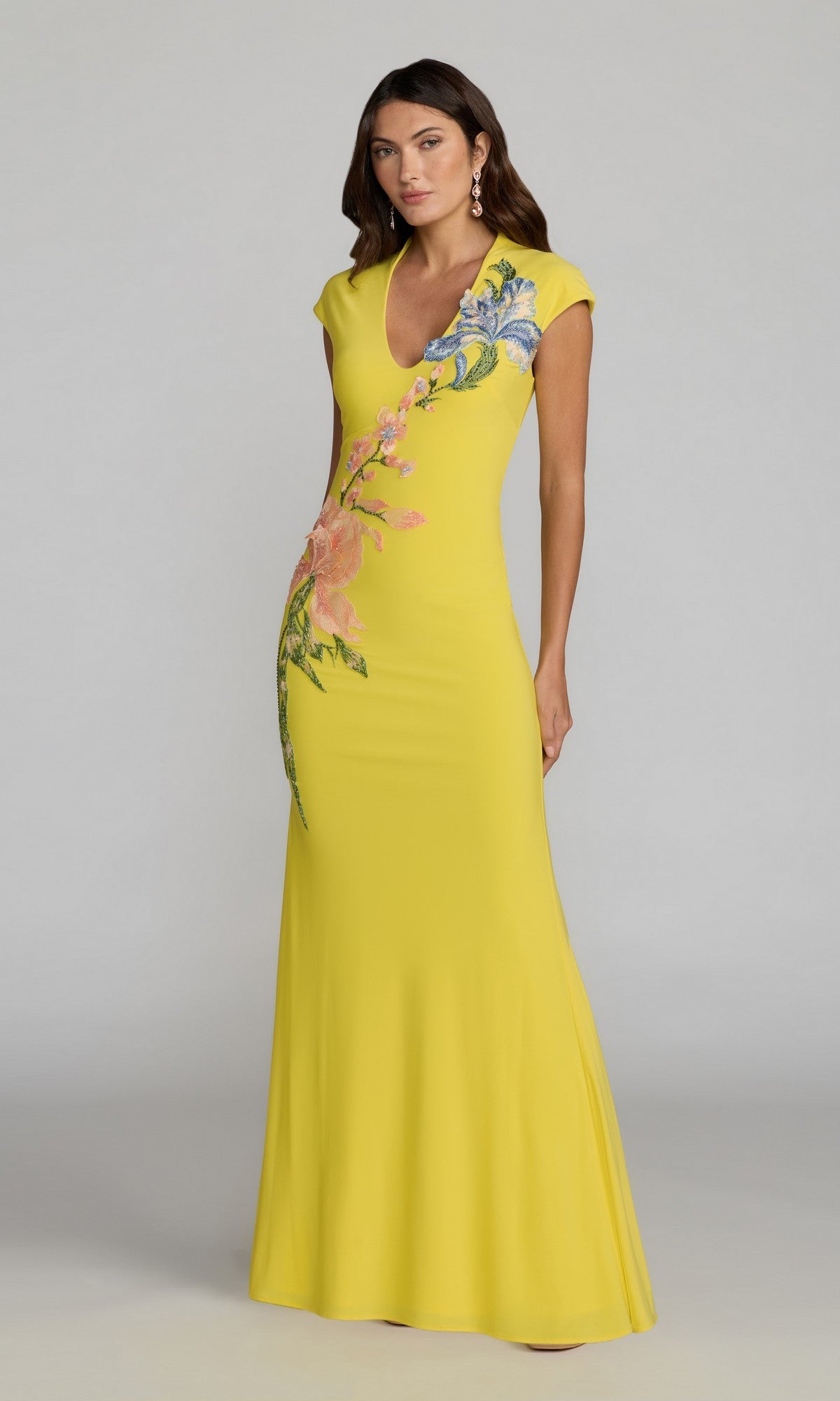 A woman stands against a plain gray background wearing the Mac Duggal 13249 long formal dress in Yellow Multi, a floor-length gown featuring short sleeves and beaded floral appliqué embellishing the upper bodice and hip.