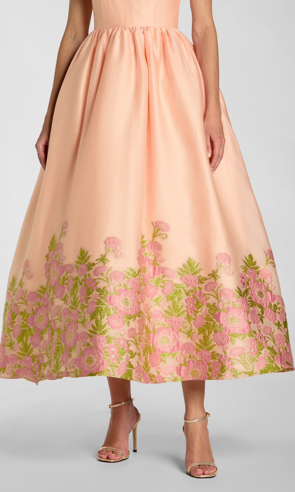 A woman wearing the Mac Duggal 13266 tea-length party dress in peach organza, featuring a sleeveless bodice and a full skirt with floral-embroidered hem in shades of pink and green; paired with strappy heels.
