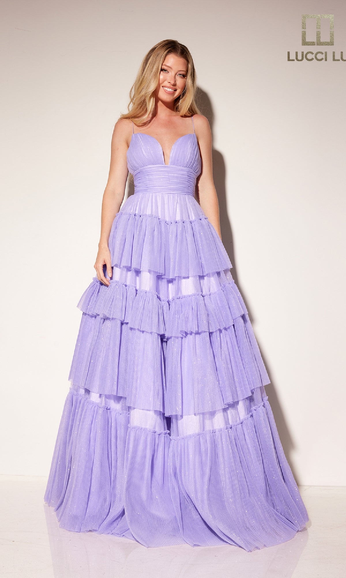 A woman stands against a plain backdrop wearing the Tiered Ruffled Glitter Prom Dress: Lucci Lu 1357, featuring lavender color, a deep V neckline, and floor-length tiered ruffles.