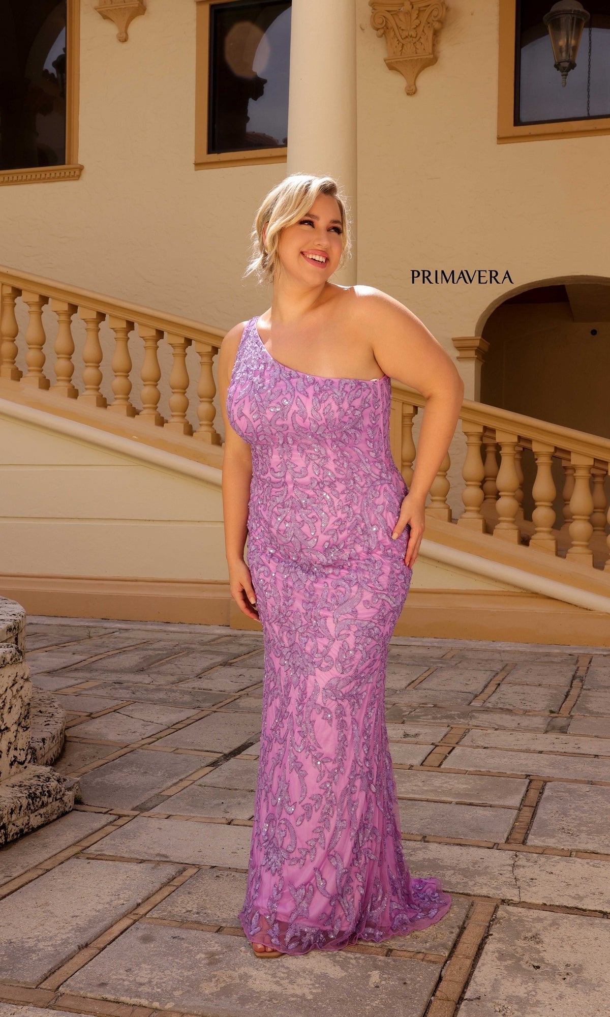 A woman wears the Primavera Plus-Size Long Beaded Prom Dress 14049 in light purple, standing outdoors before a cream building and staircase, highlighting the gown’s designer elegance.