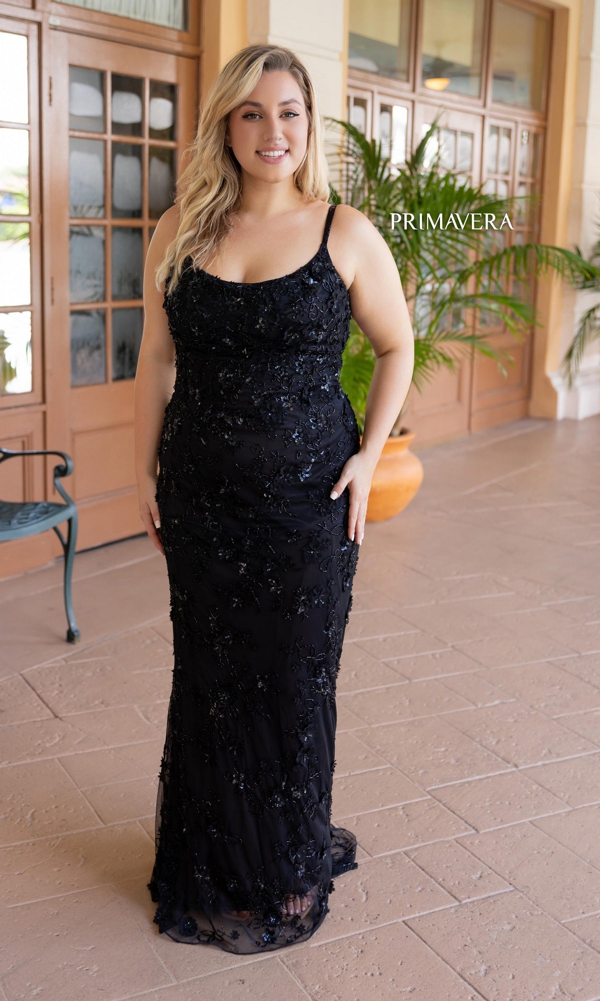 A woman in the Plus-Size Long Beaded Primavera Prom Dress 14050 stands on a tiled patio before wooden doors and a potted plant.