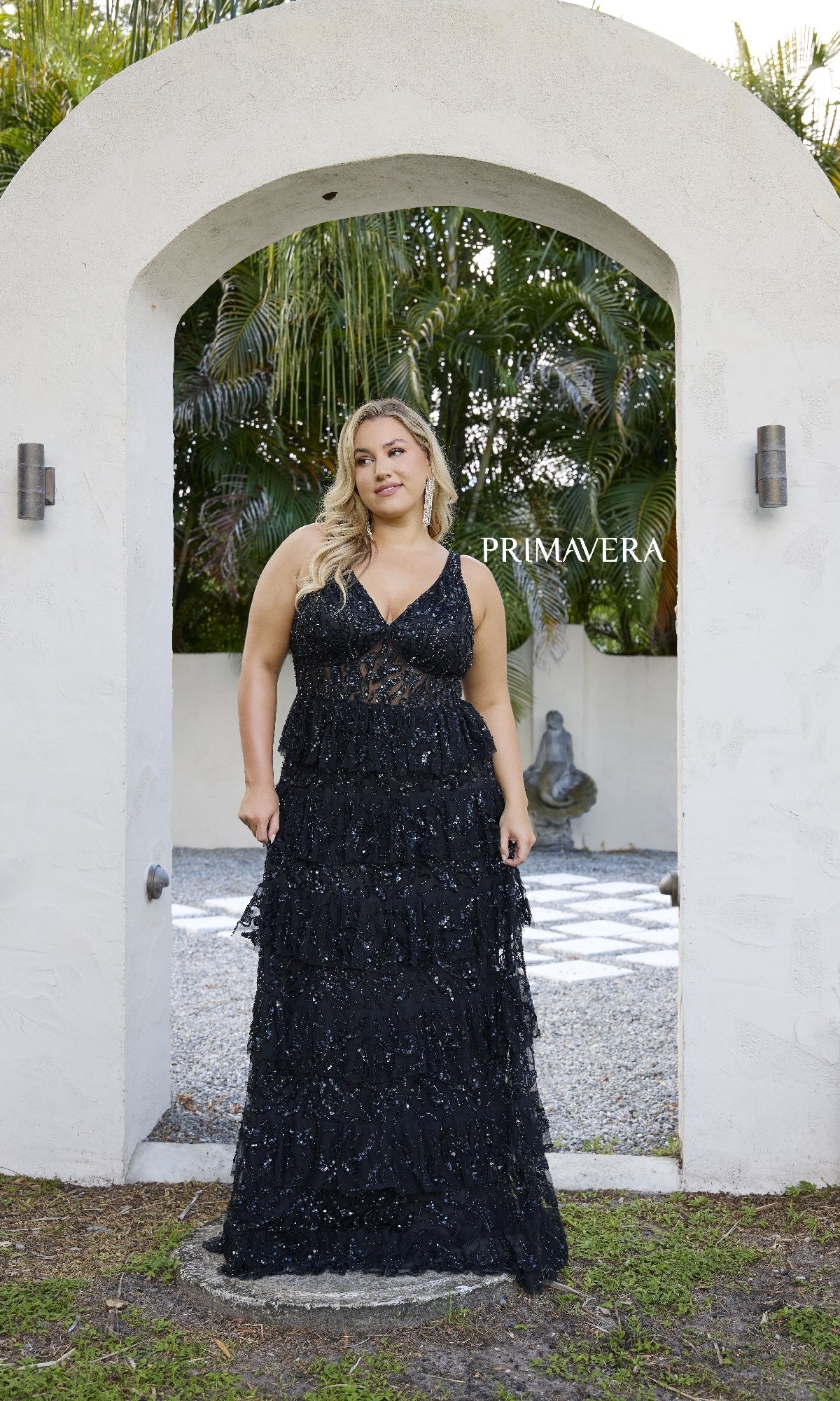 A woman wears the Primavera 14073 Long Plus-Size Prom Dress, a sleeveless black sequin evening gown, standing outdoors under a white stone archway with greenery and a graceful statue in the background.