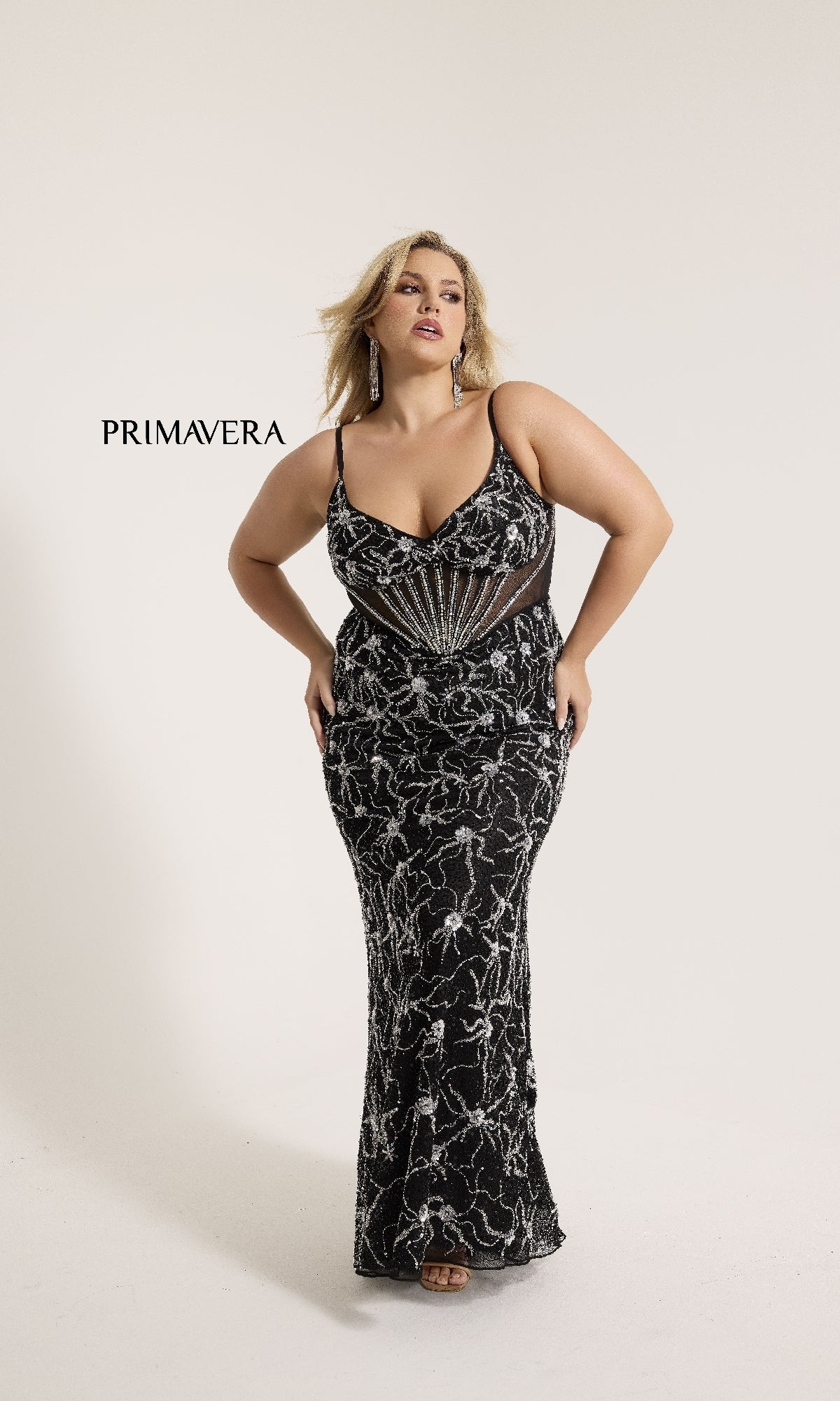 A woman models the Primavera 14079, a plus-size long sequin prom gown with black and silver beading and sheer panels, standing against a plain white background.