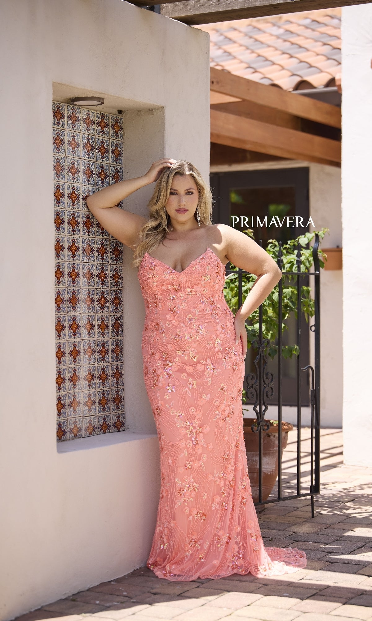 A woman models the Long Plus-Size Prom Dress, Primavera 14080—a peach, strapless gown with floral embellishments—posing outdoors by a white wall and patterned tile. The word