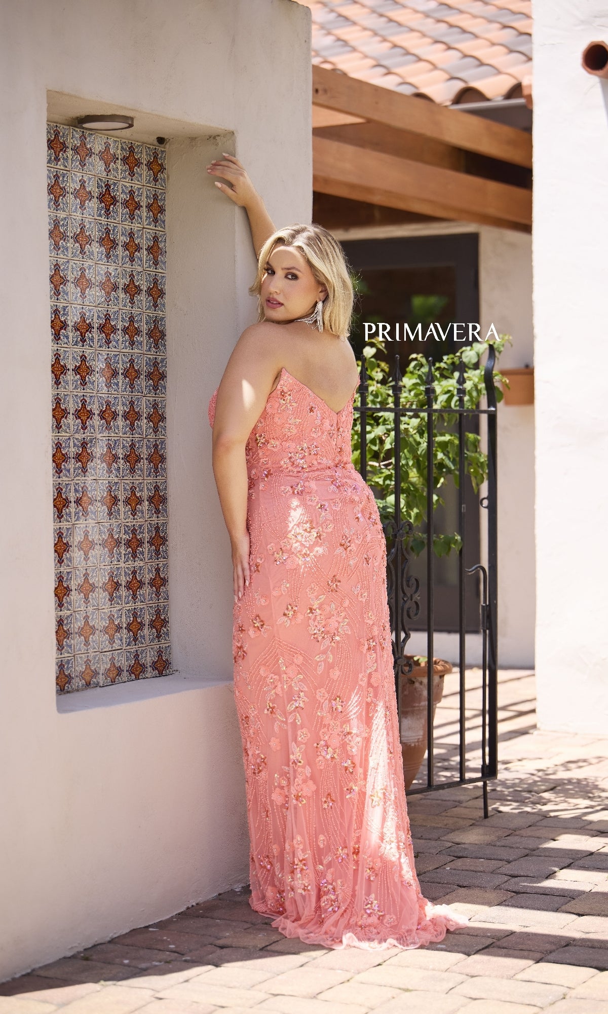 A woman wearing the Long Plus-Size Prom Dress: Primavera 14080, with pink floral details, stands outdoors facing away and looking over her shoulder, as