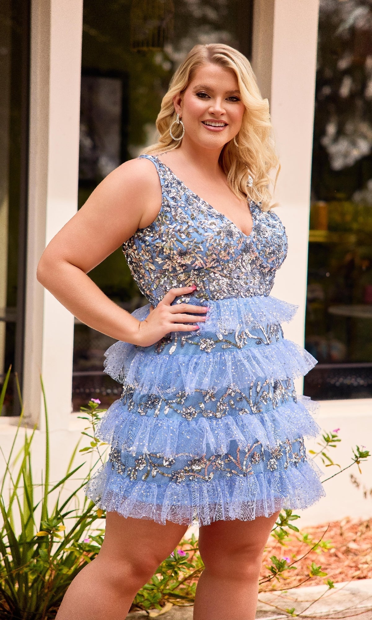 A confident blonde woman smiles outside in front of a window, wearing the Primavera 14092 Plus-Size Short Beaded Hoco Dress—a blue sleeveless dress with ruffles and silver embellishments.