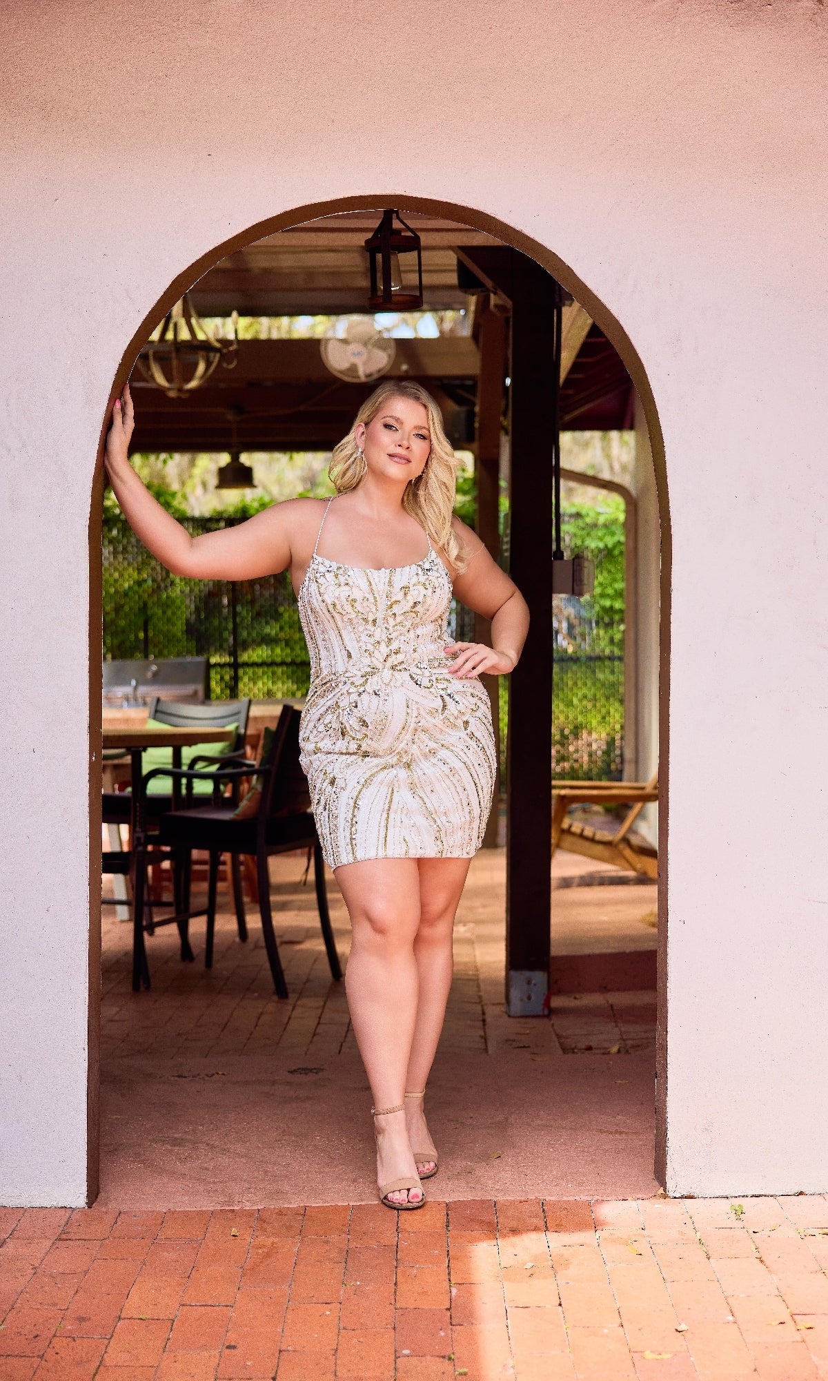 A woman wearing the Plus-Size Mini Beaded Hoco Dress: Primavera 14093 stands confidently under an arched doorway, her hand on the arch, with outdoor seating visible in the background.