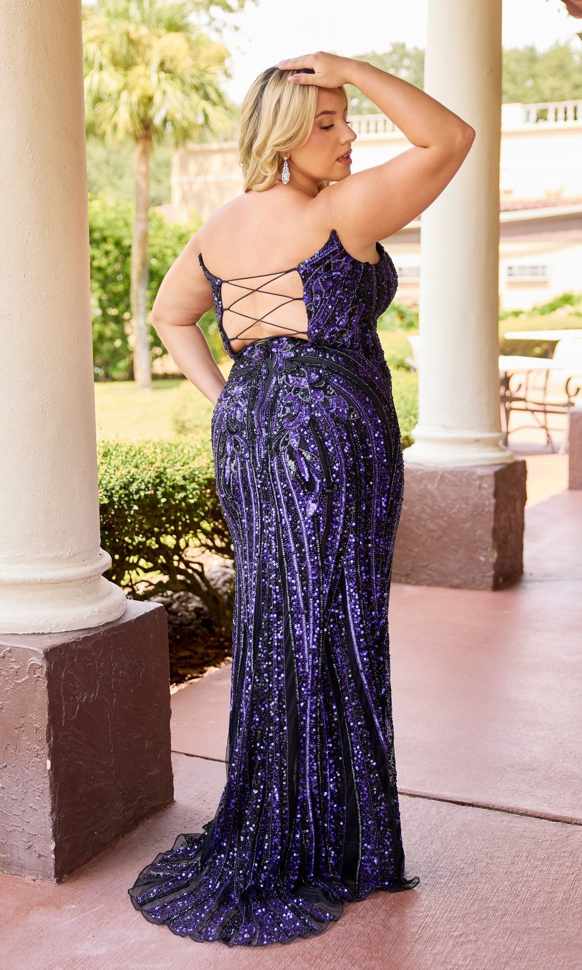 A woman in the Long Plus-Size Prom Dress Primavera 14103, featuring purple sequins and a lace-up back, stands outdoors near columns facing away from the camera.