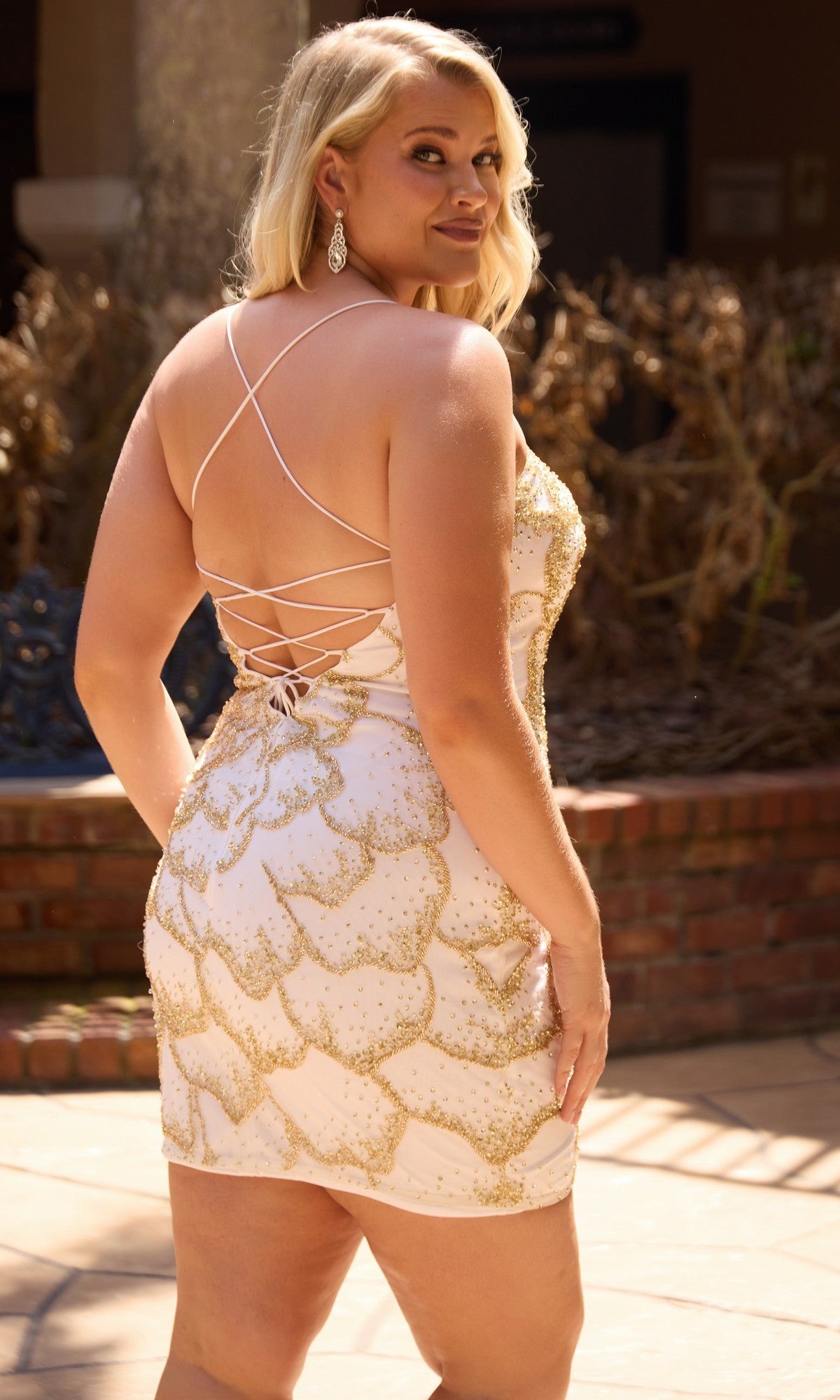 A woman poses outdoors in front of a brick planter with dry plants, wearing the Primavera 14115 Plus-Size Homecoming Dress—a short, light-colored mini dress featuring gold details, crystal beading, and a lace-up back.