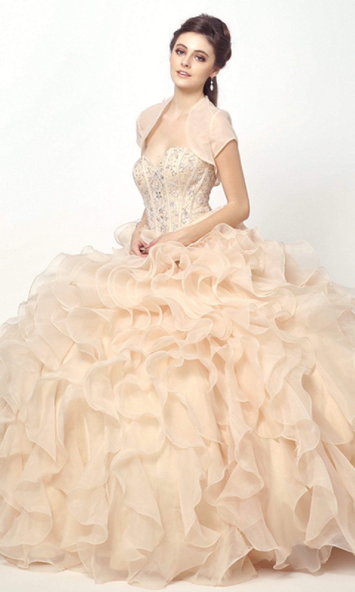 A woman poses elegantly against a plain white background in the Champagne Quinceanera Dress 1418 By Juliet, featuring a strapless beaded corset bodice and a voluminous, ruffled pale peach skirt.