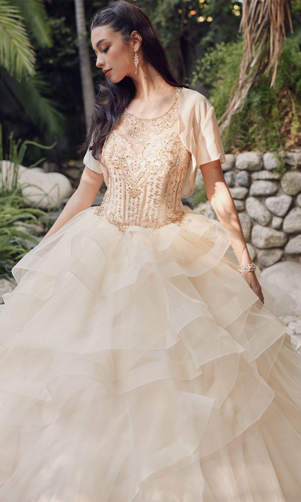 A woman stands outdoors in the Quinceanera Dress By Juliet 1423, featuring a beaded high neck bodice, short sleeves, and a layered tulle skirt, surrounded by stone and greenery.