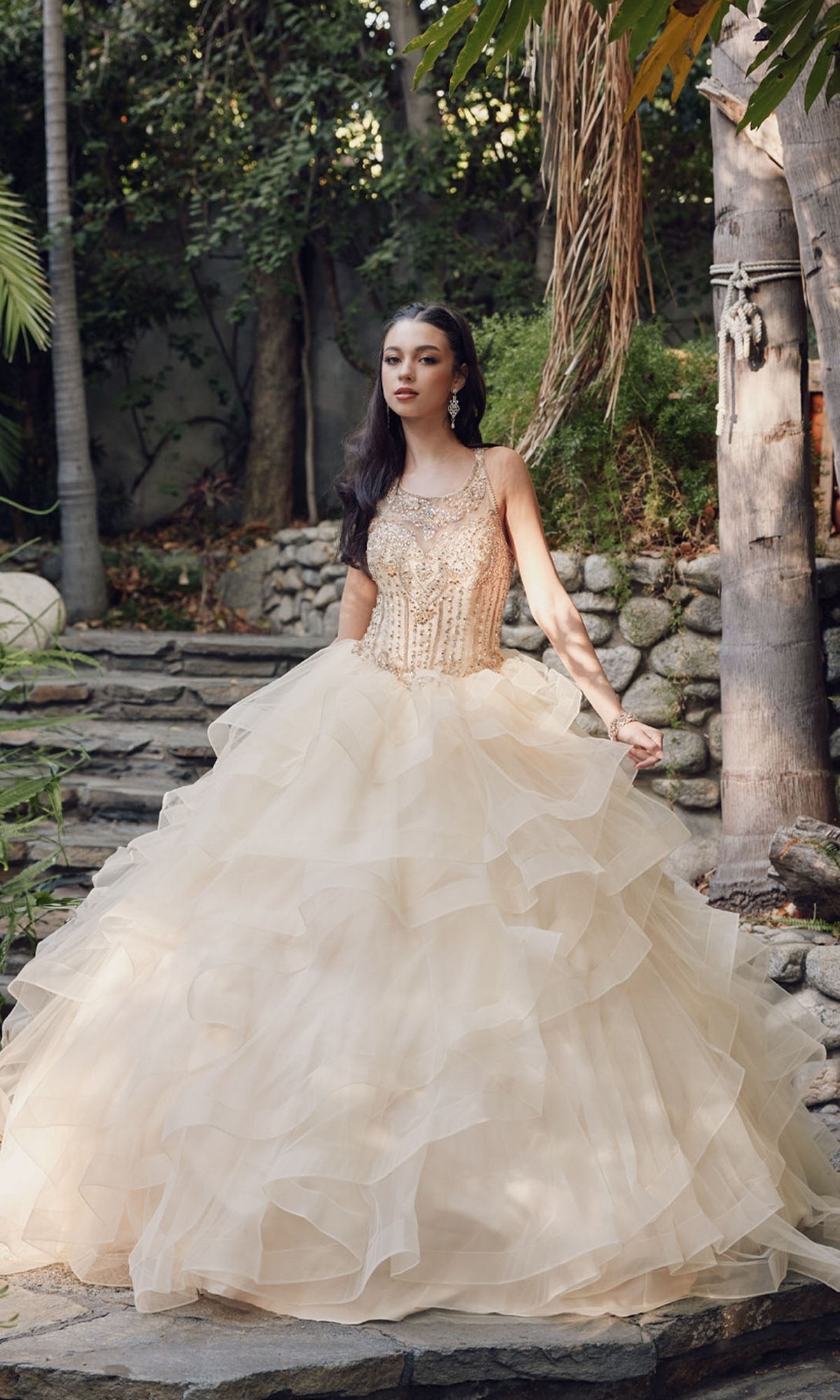 A woman stands on stone steps outdoors wearing the elegant Quinceanera Dress By Juliet 1423, featuring a cream hue, beaded high neck bodice, and layered tulle skirt. Trees and lush garden greenery provide a stunning backdrop.