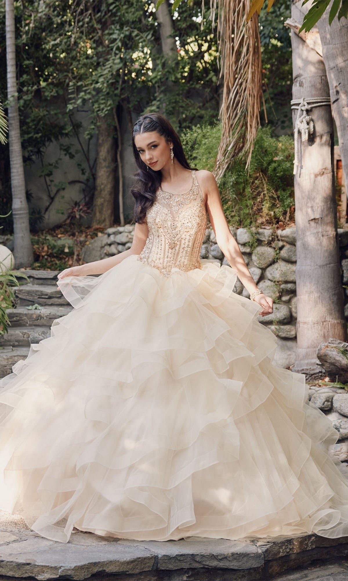 A woman in the Quinceanera Dress By Juliet 1423, a sleeveless layered A-line gown with a beaded high neck bodice, stands outdoors on stone steps amid greenery and trees.