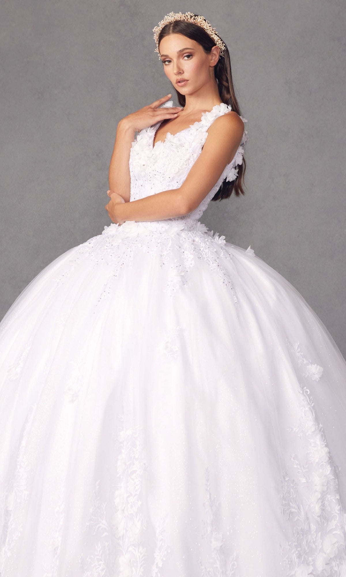 A woman stands against a gray background wearing the Juliet 1437W Traditional White Quinceañera Dress, featuring floral details and a crown.