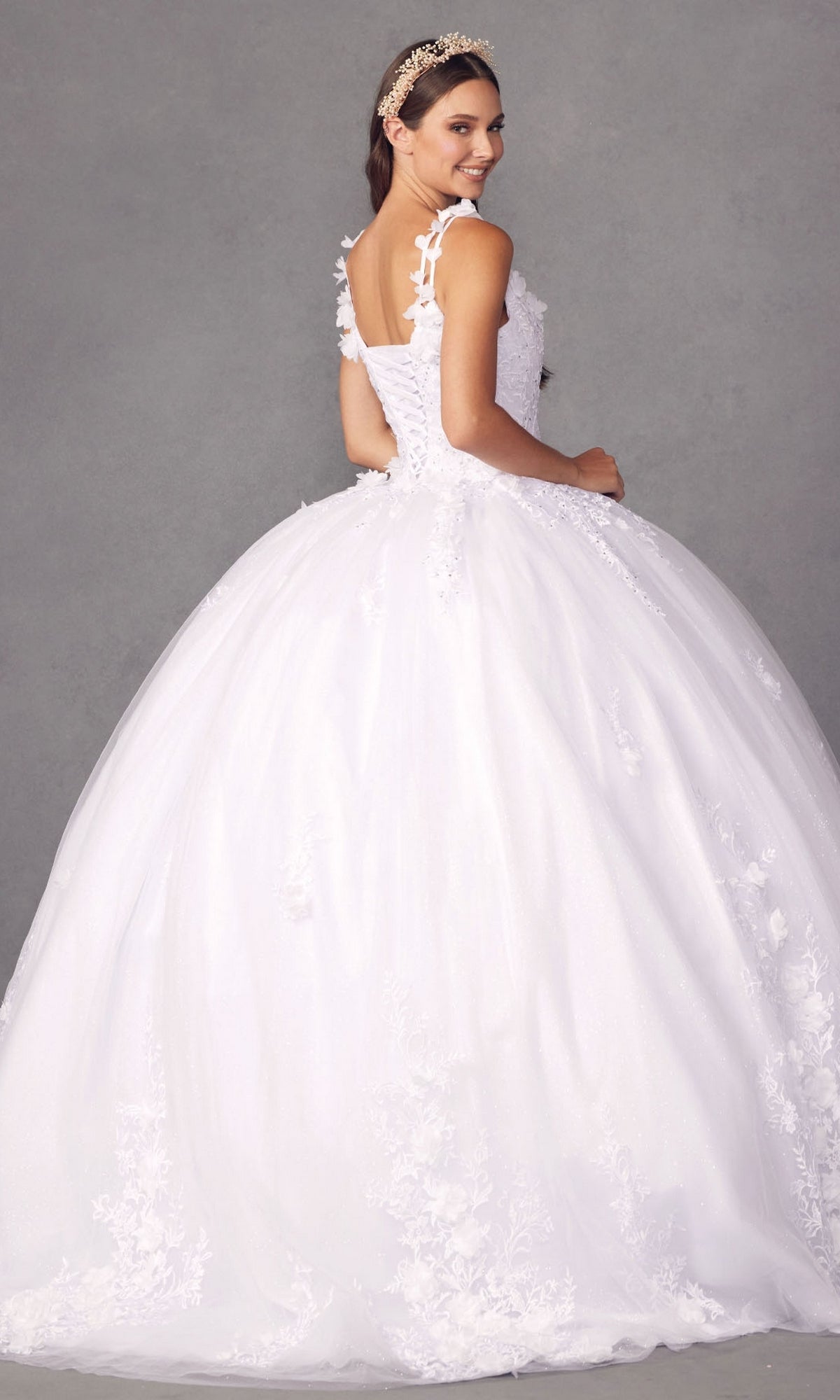 A woman wearing the Juliet 1437W Traditional White Quinceañera Dress with floral appliqués and a decorative headband stands against a plain gray background, looking over her shoulder.