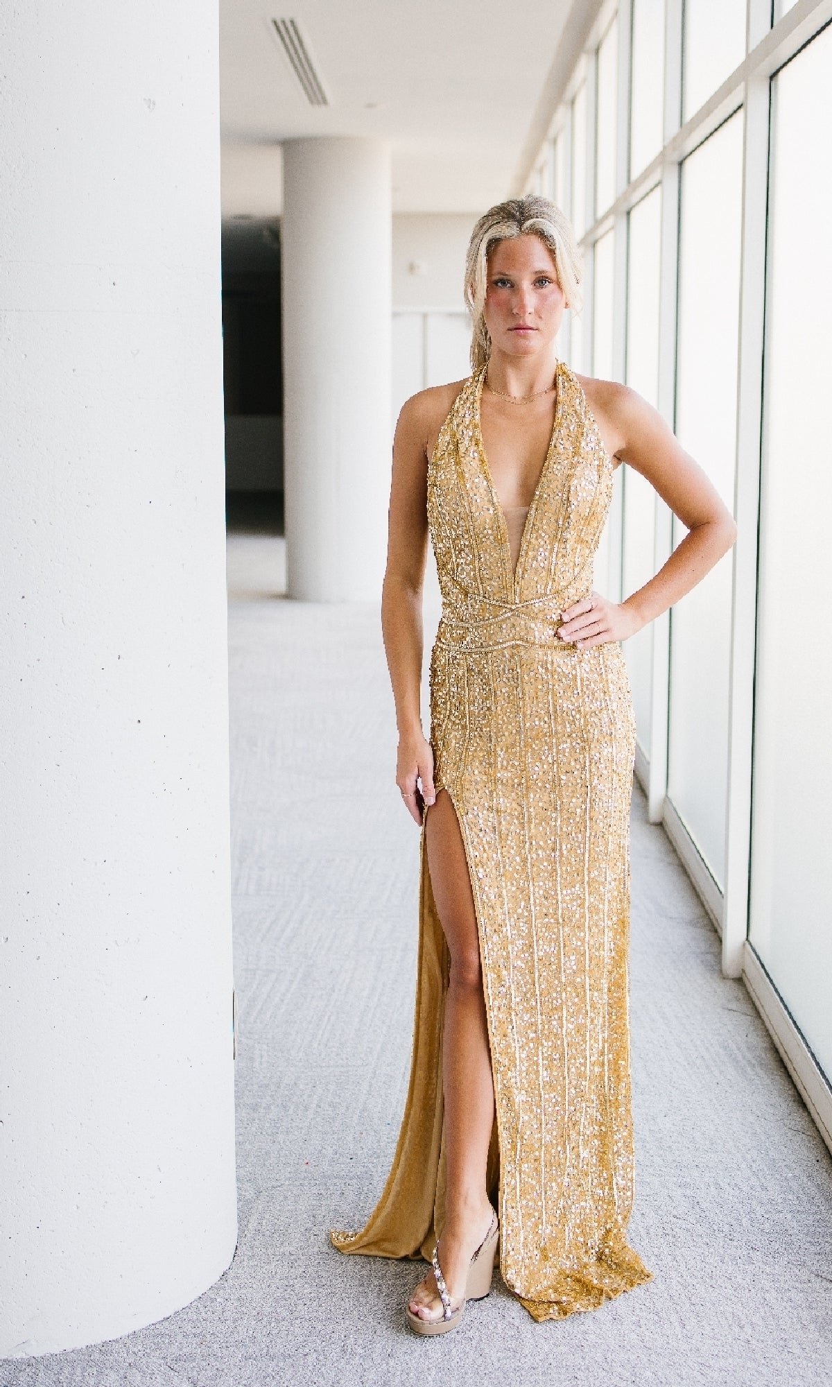 A woman poses elegantly indoors before large windows, wearing the Aleta 1441 Backless Long Sequin Halter Prom Dress—sleeveless with gold sequins, high slit, and open back—paired with platform heels, one hand on her hip.