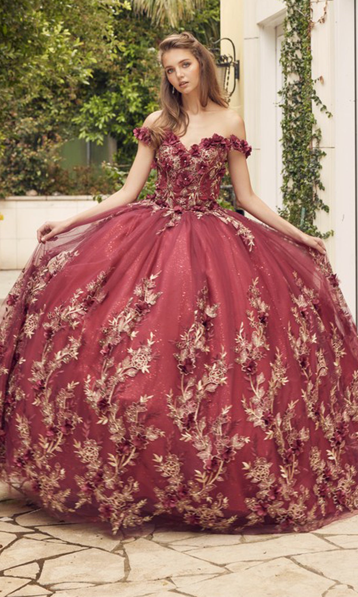 A young woman poses outdoors in the Juliet 1447 Off-the-Shoulder Quinceañera Dress, a burgundy glitter ball gown with gold beaded embroidery and a full sparkling tulle skirt.