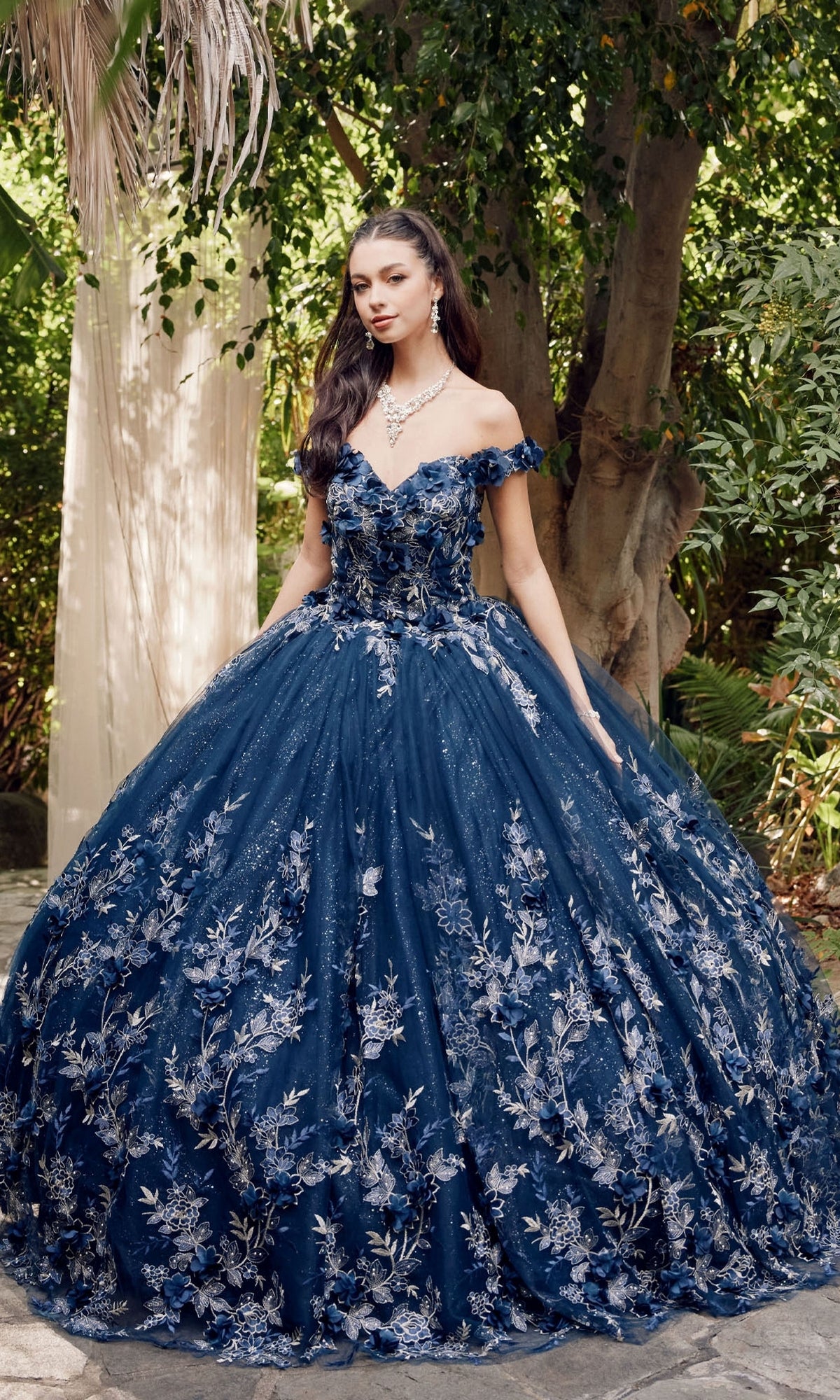 A woman stands outdoors in the Juliet 1447 Off-the-Shoulder Quinceañera Dress, a navy blue glitter ball gown with silver beaded embroidery, surrounded by lush greenery.