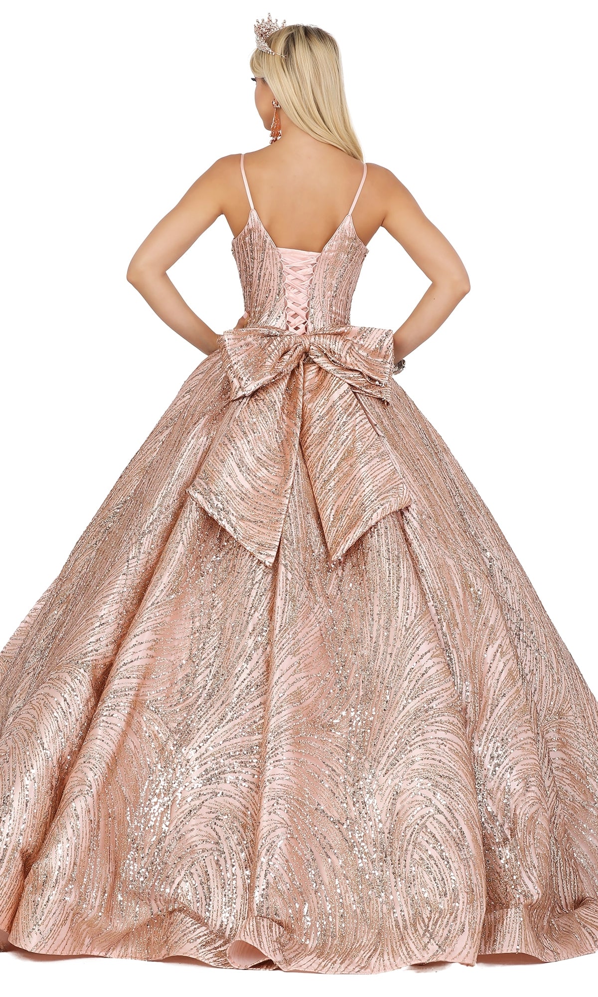 Wearing Quinceanera Dress 1447 by Dancing Queen, a woman in a pink, glittery A-line gown with a large back bow and tiara shows off delicate lace embroidery as she stands with her back to the camera.