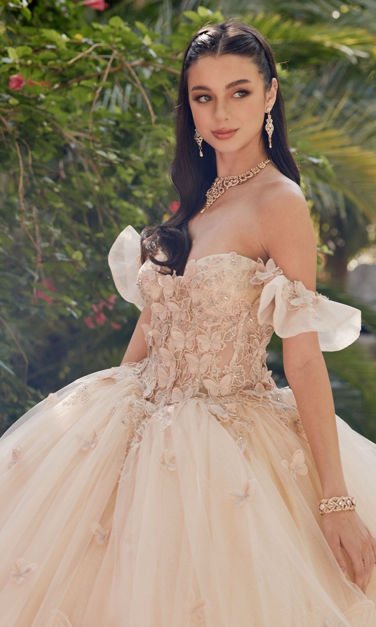 A woman stands outdoors among green foliage, wearing the Quinceanera Dress By Juliet JT1448J—an off-the-shoulder, pastel pink gown with floral appliqué—paired with matching jewelry and her long, wavy hair.