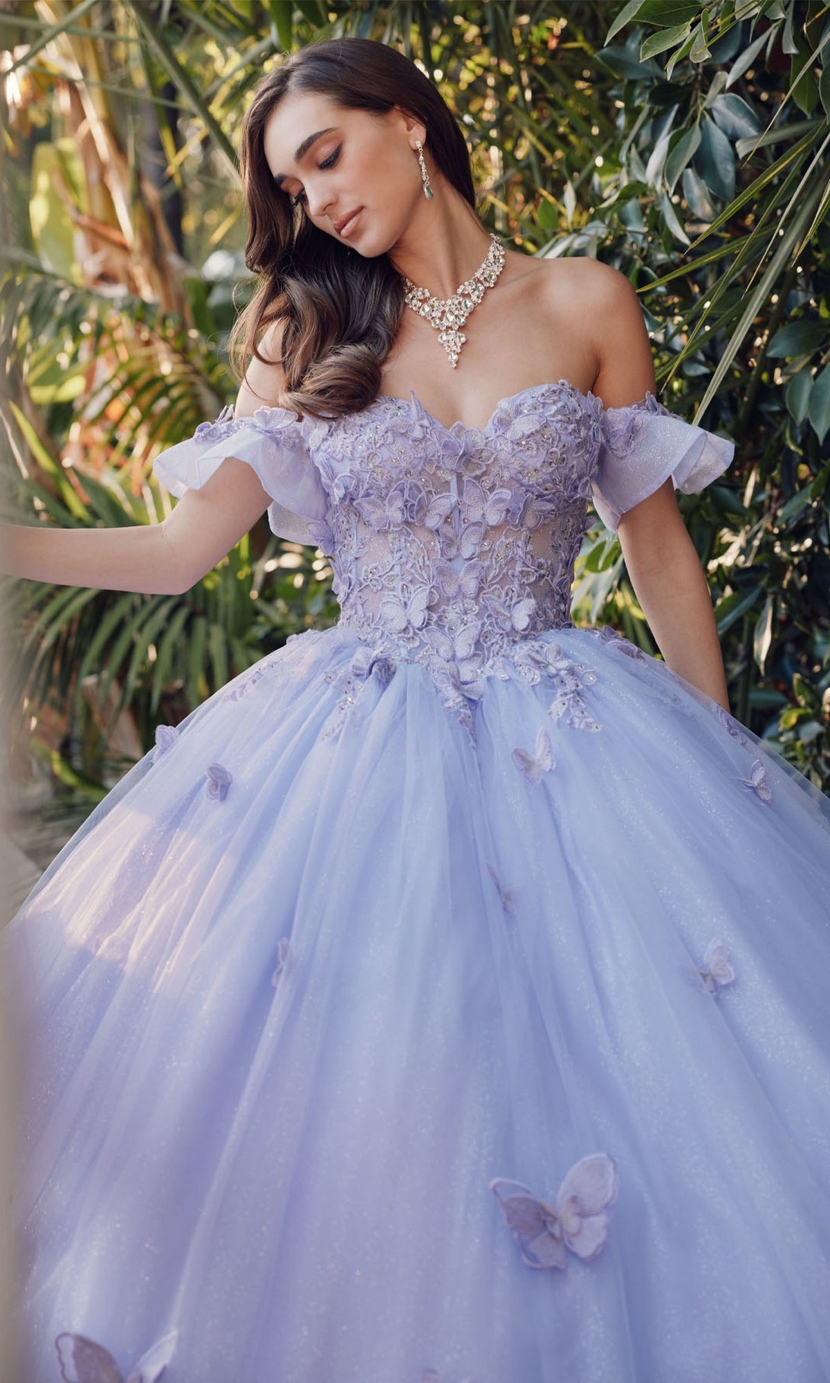 A woman stands outdoors among green plants wearing the Quinceanera Dress By Juliet JT1448J, a lavender off-the-shoulder ballgown with floral and butterfly details, paired with a necklace and earrings.