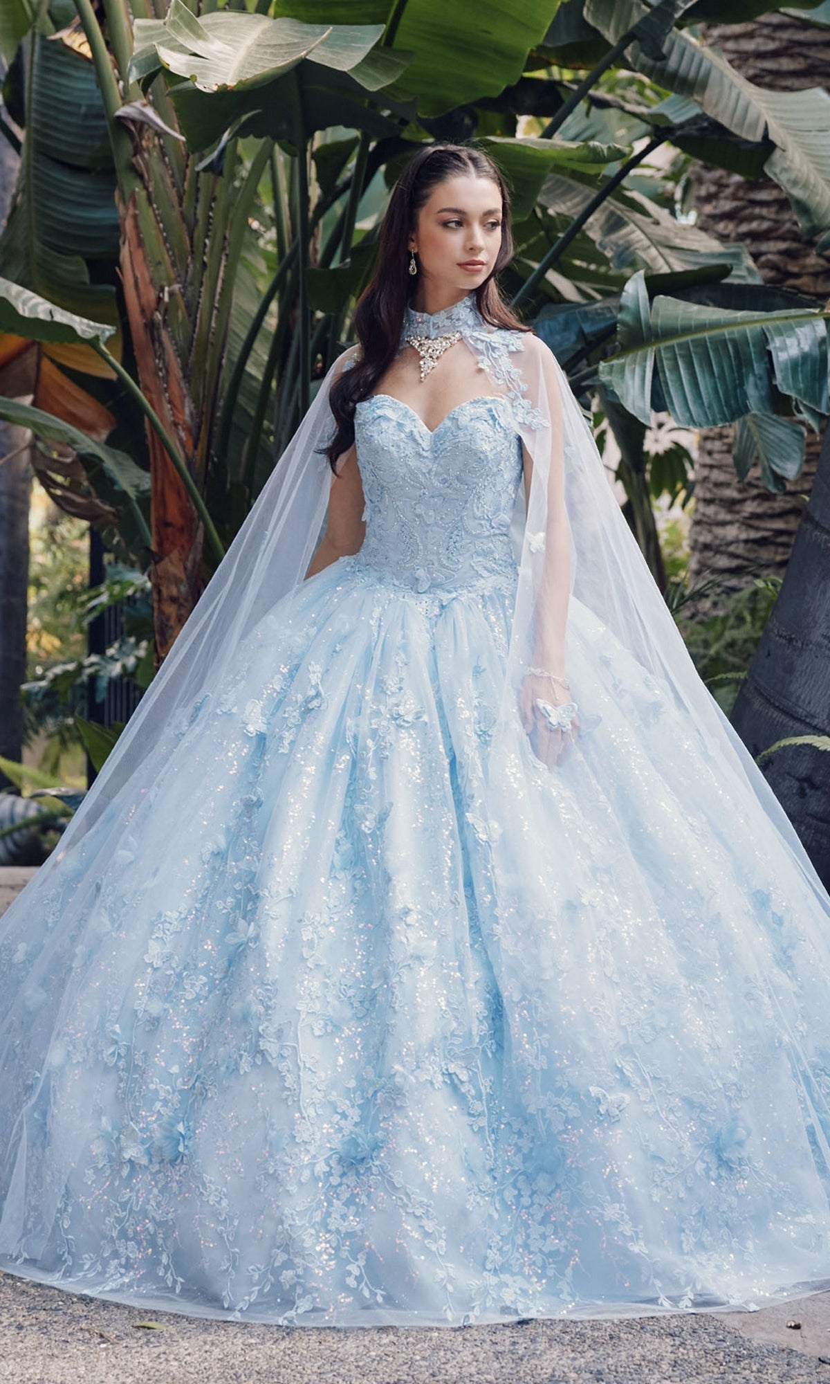 A woman stands outdoors in the Quinceanera Dress JT1449J by Juliet, a Bahama blue strapless sweetheart ball gown with sheer cape and floral details, surrounded by lush green tropical plants.
