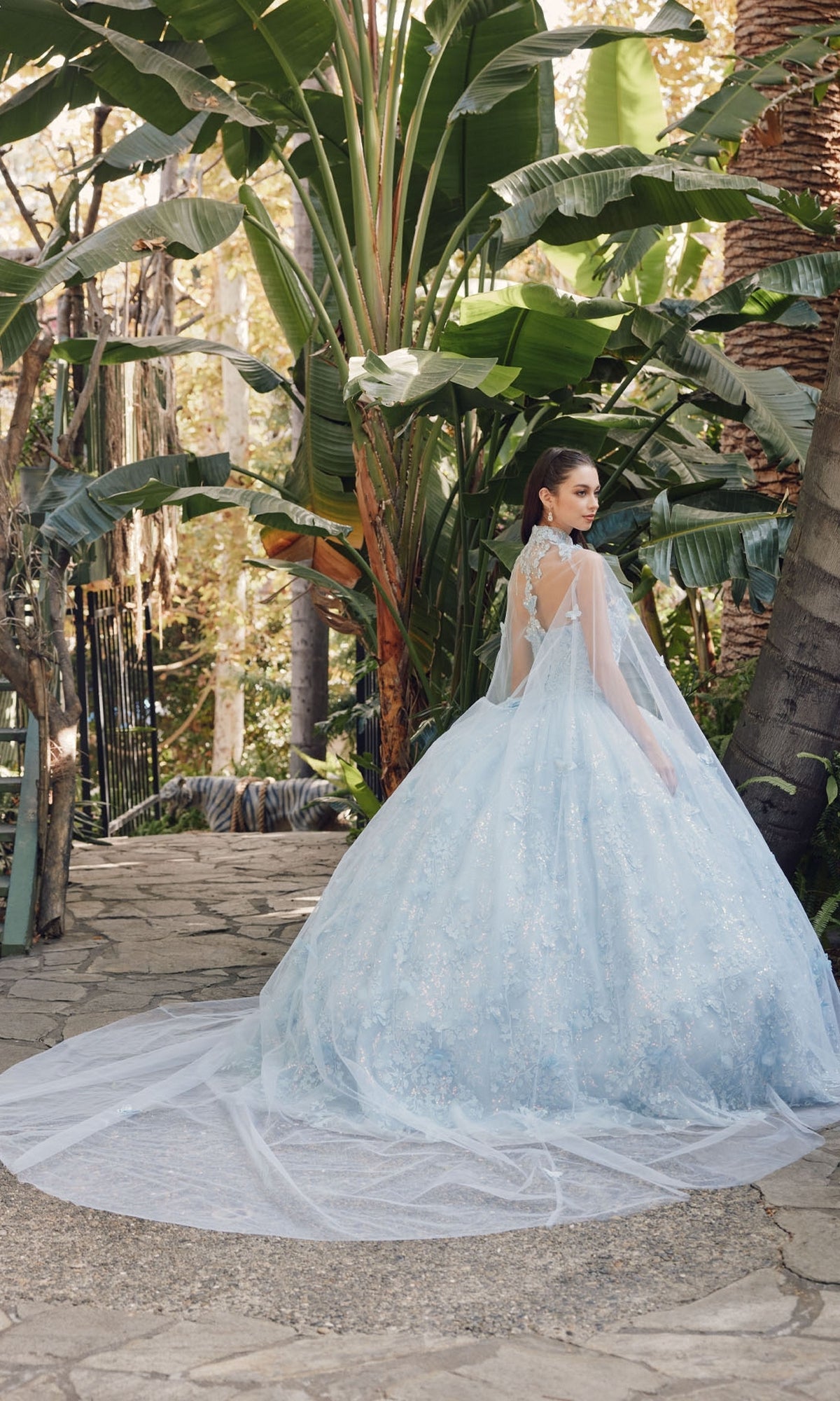 A woman in the Quinceanera Dress JT1449J by Juliet, featuring a Bahama blue strapless sweetheart neckline and a-line ball gown, stands on a stone path surrounded by tall tropical plants and trees.
