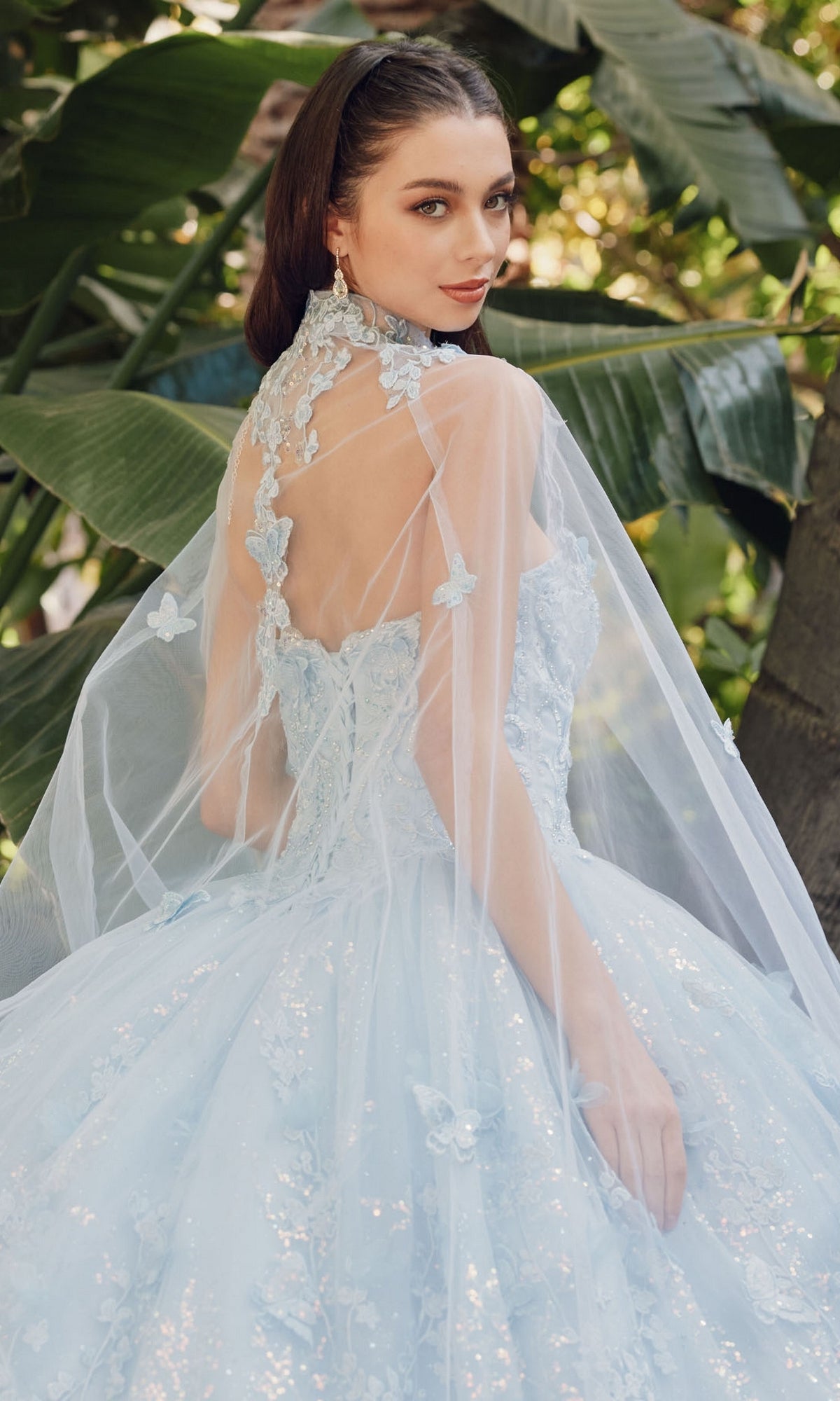 A woman wears the Quinceanera Dress JT1449J by Juliet in Bahama blue, featuring floral lace, a sheer cape, strapless sweetheart neckline, and A-line ball gown silhouette as she stands outdoors amidst lush green leaves.