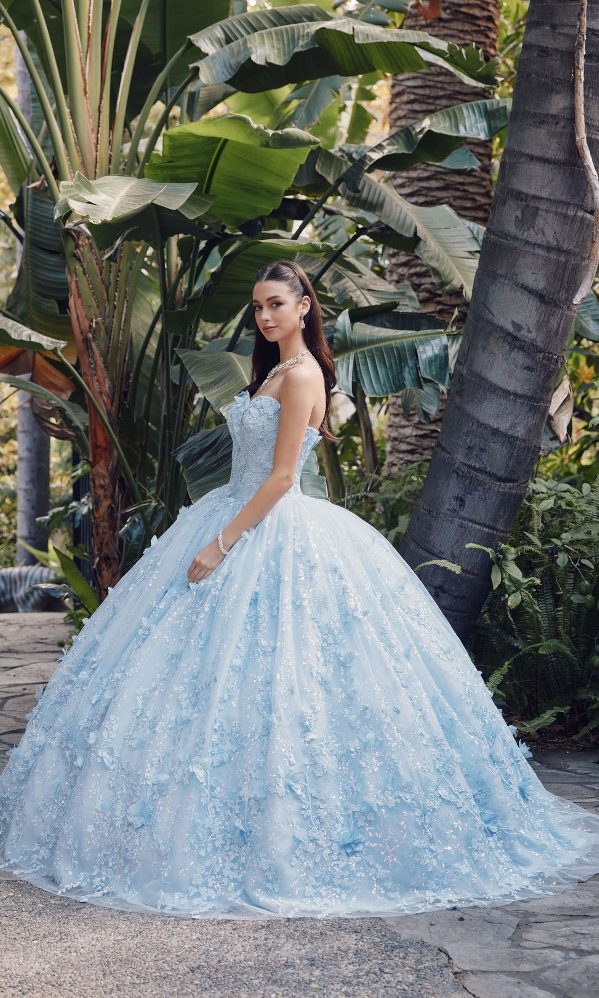 A young woman in the Quinceanera Dress JT1449J by Juliet, featuring a Bahama blue strapless sweetheart neckline, stands outdoors on a stone path surrounded by lush tropical plants and trees.