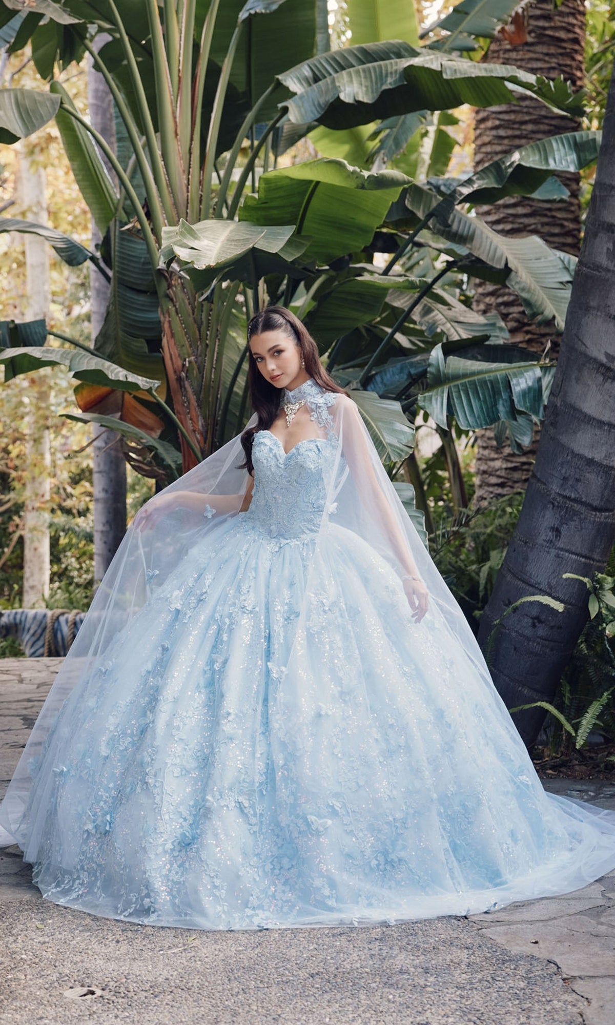 A woman stands outdoors before tall tropical plants, wearing the JT1449J Quinceanera Dress by Juliet in Bahama blue, featuring floral details, lace, a sheer cape, and an elegant strapless sweetheart neckline.