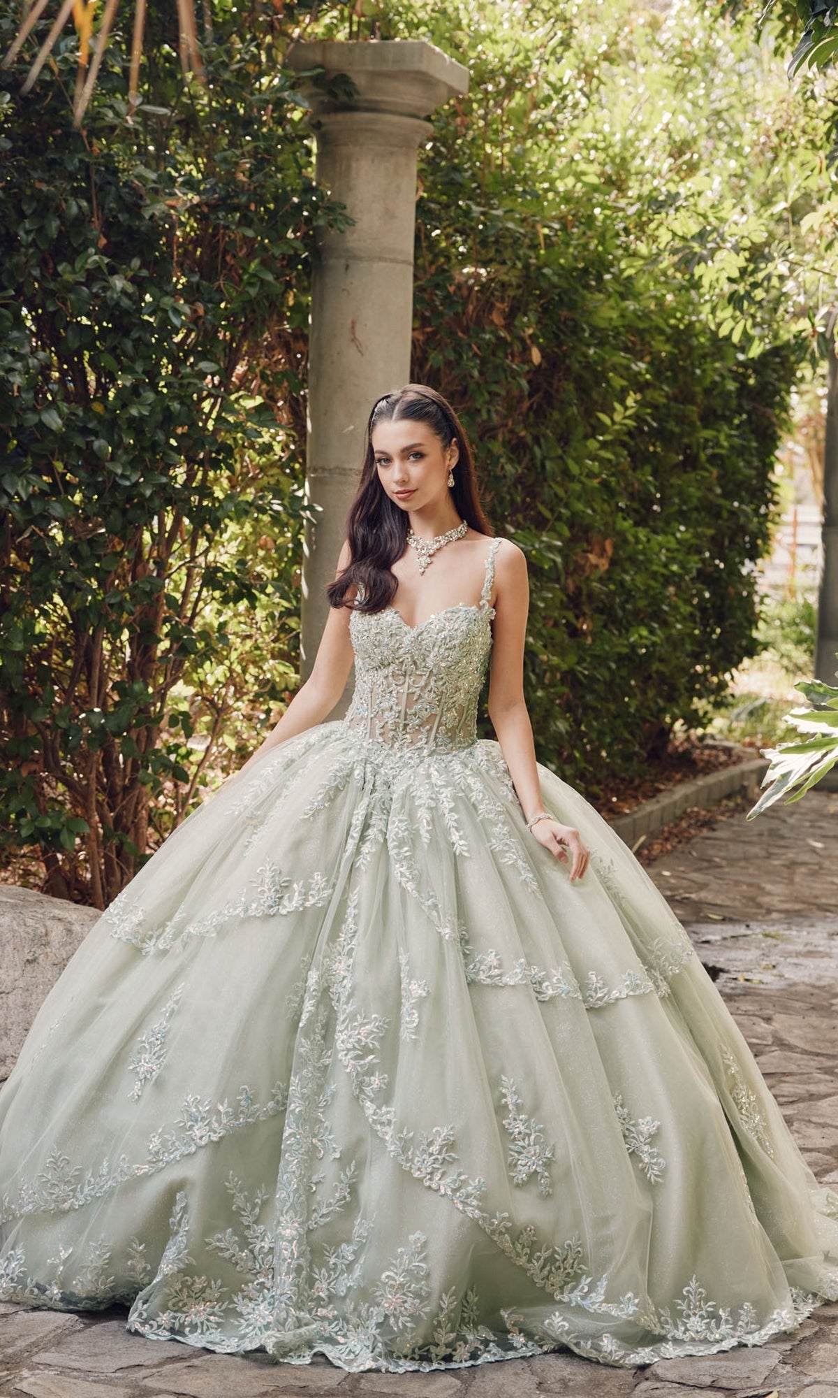 A woman stands outdoors in the Quinceanera Dress By Juliet JT1450J, a pale green ball gown with a sweetheart neckline and intricate tulle embroidery, framed by lush greenery and stone columns.
