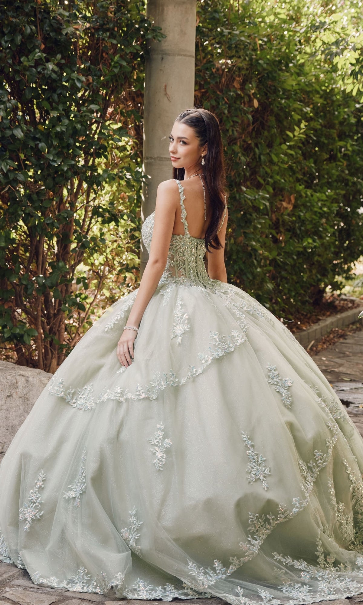 Wearing the Quinceanera Dress By Juliet JT1450J, a woman stands outdoors in greenery and on stone pathways, looking back over her shoulder in a light green ball gown with floral embroidery.
