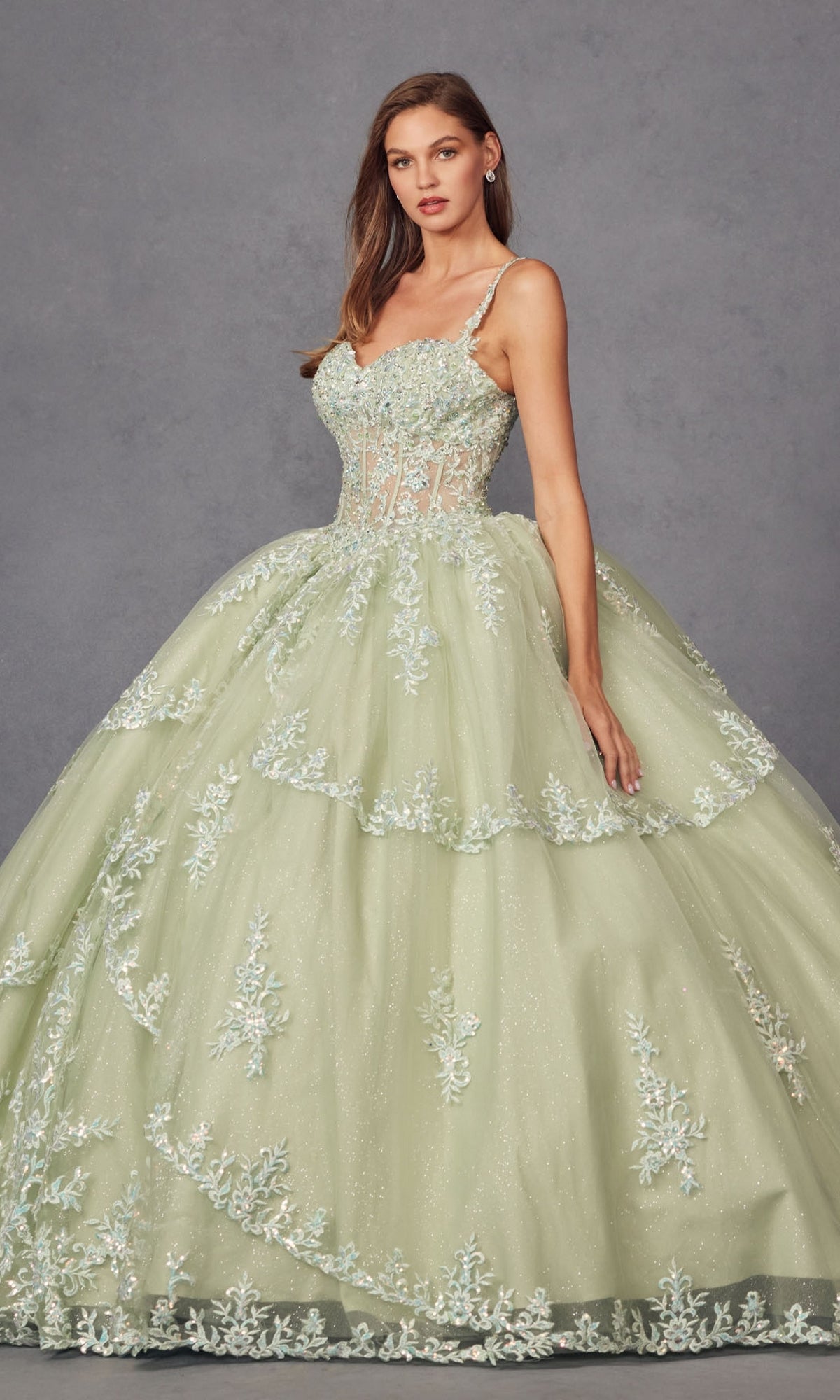 A woman stands against a plain gray background wearing the Quinceanera Dress By Juliet JT1450J, a light green ball gown featuring floral embroidery, a fitted bodice with sweetheart neckline, and a full layered skirt.
