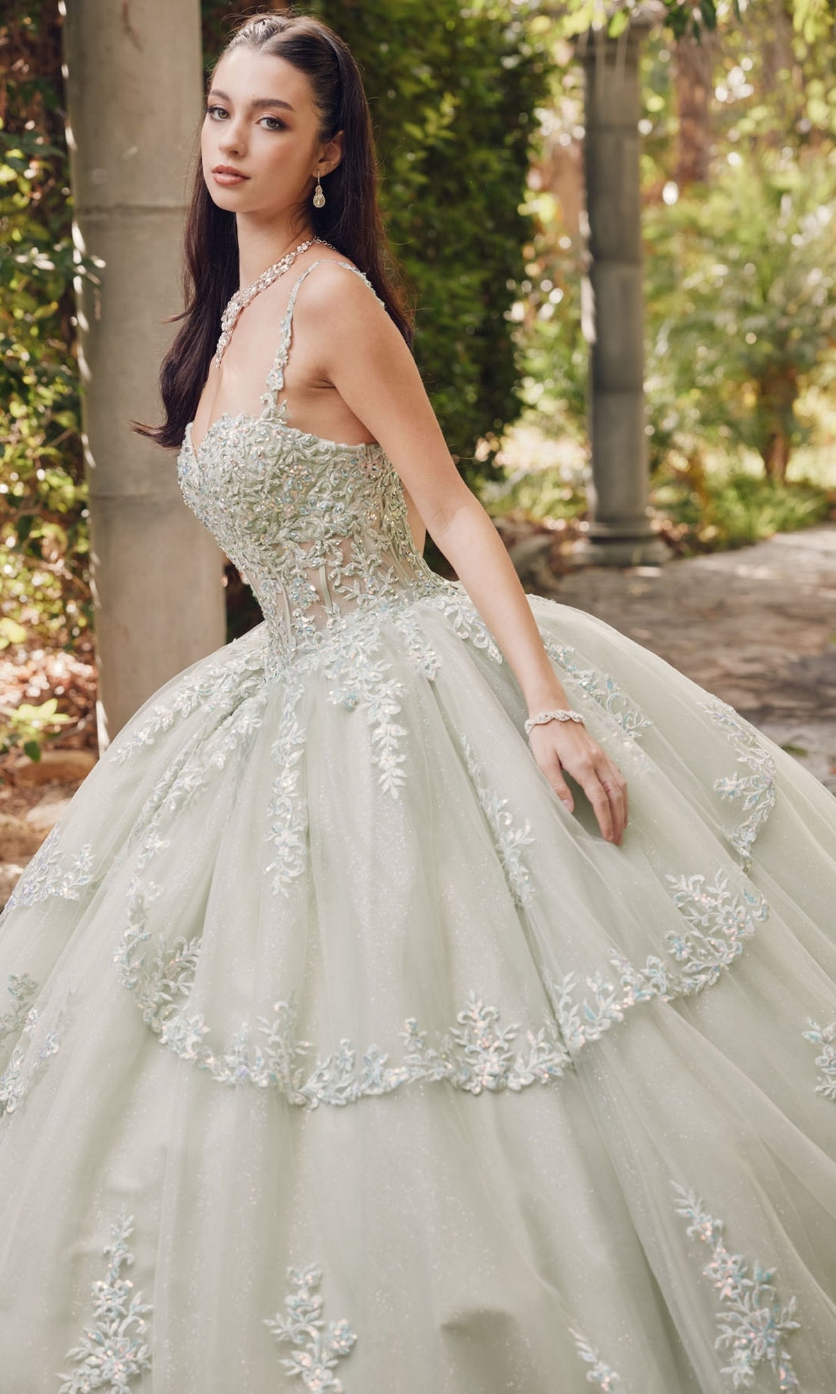 In a formal outdoor setting, a woman wears the Quinceanera Dress By Juliet JT1450J, featuring a light hue, sweetheart neckline, floral embroidered tulle with beadwork, and matching jewelry.