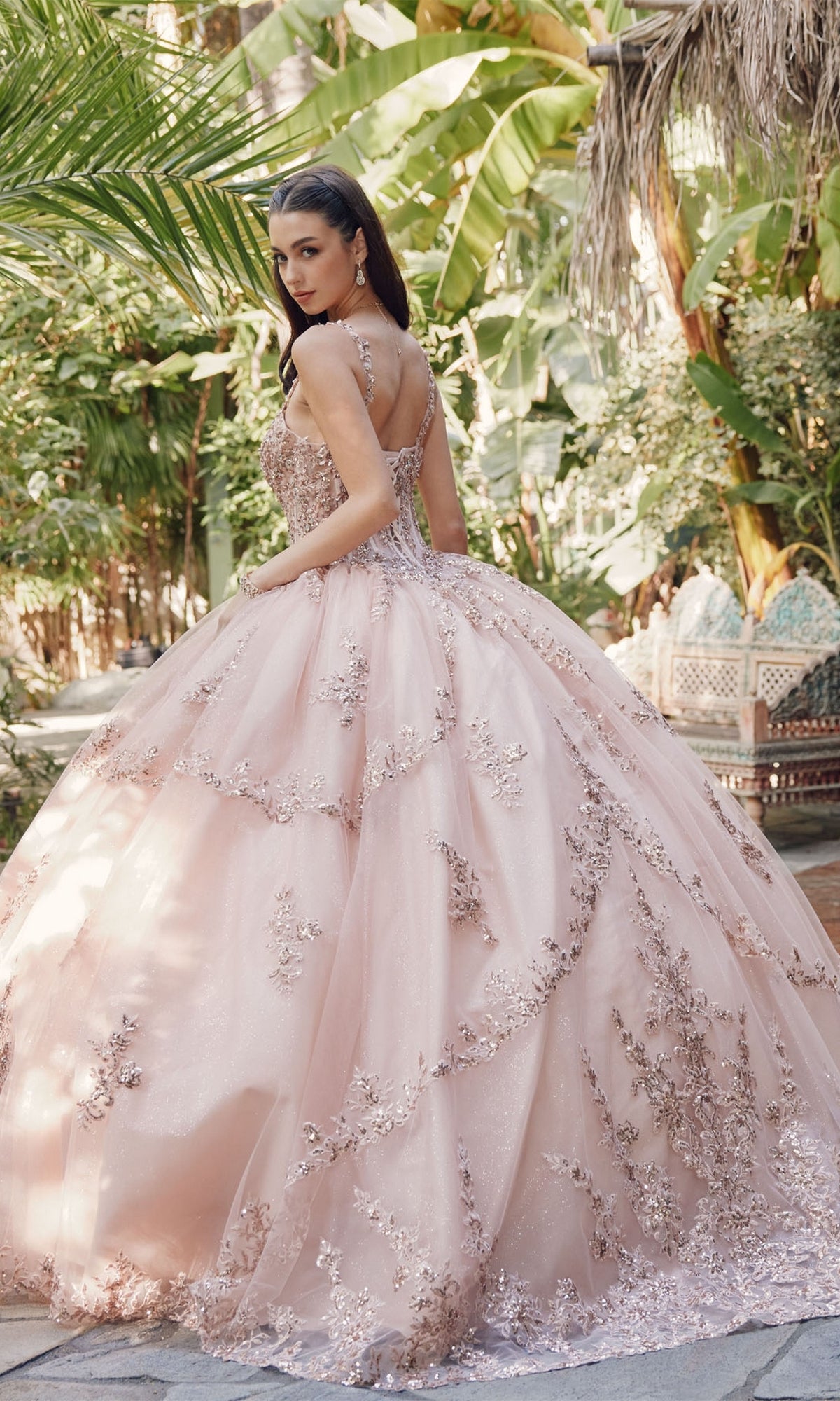 A woman in the Juliet JT1450J Quinceanera Dress, featuring a pale pink ball gown and sweetheart neckline, stands outdoors among lush greenery, glancing over her shoulder.