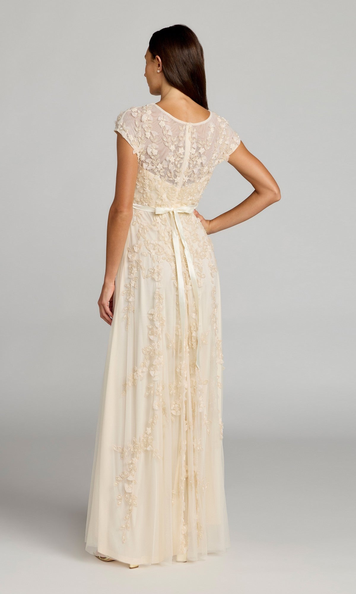 A woman stands facing away, wearing the Mac Duggal 14521 Cap-Sleeve Long Formal Dress in cream, featuring floral lace accents and a ribbon waist tie.