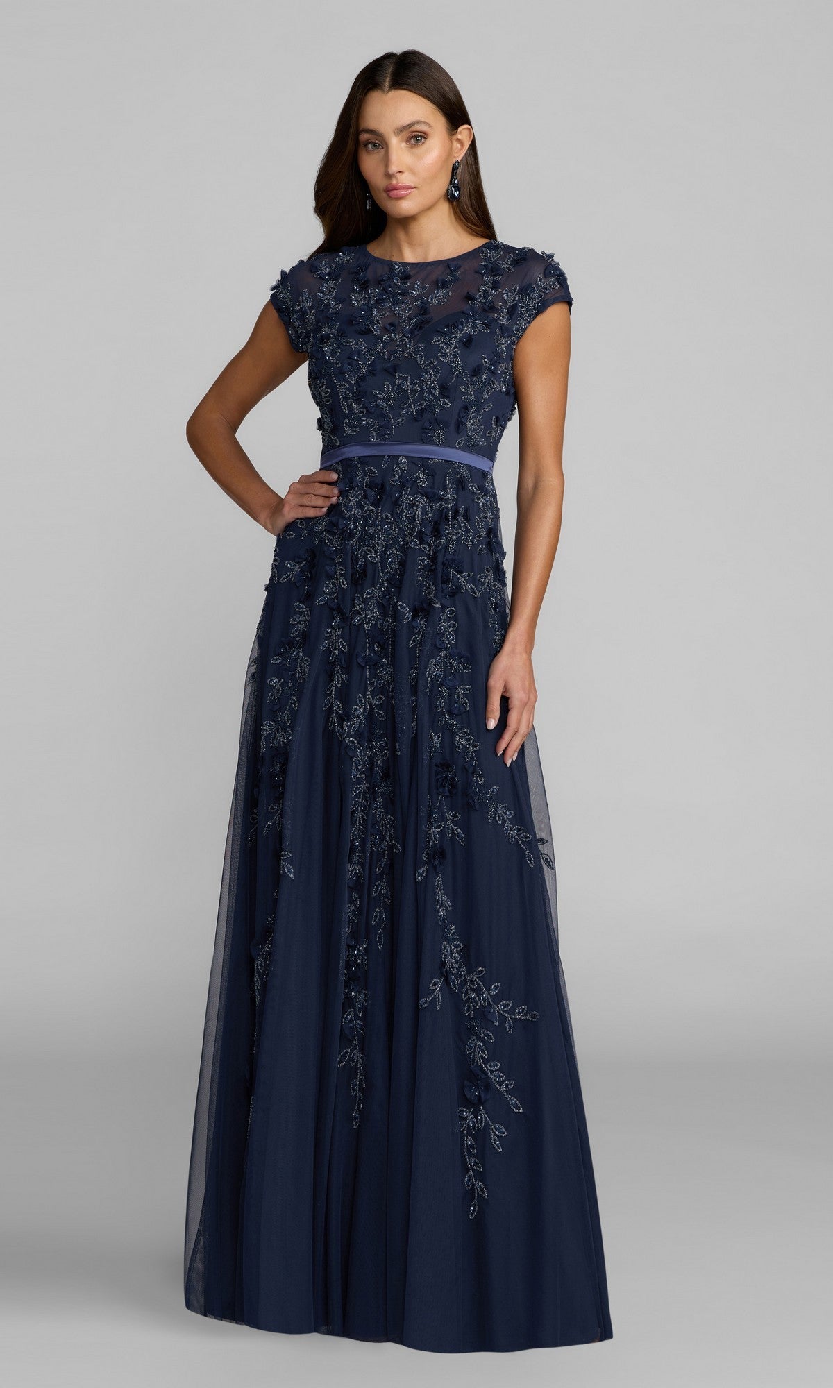 A woman models the Mac Duggal 14521 Cap-Sleeve Long Formal Dress, a floor-length navy gown with floral embroidery and a fitted waistband, set against a plain gray background.