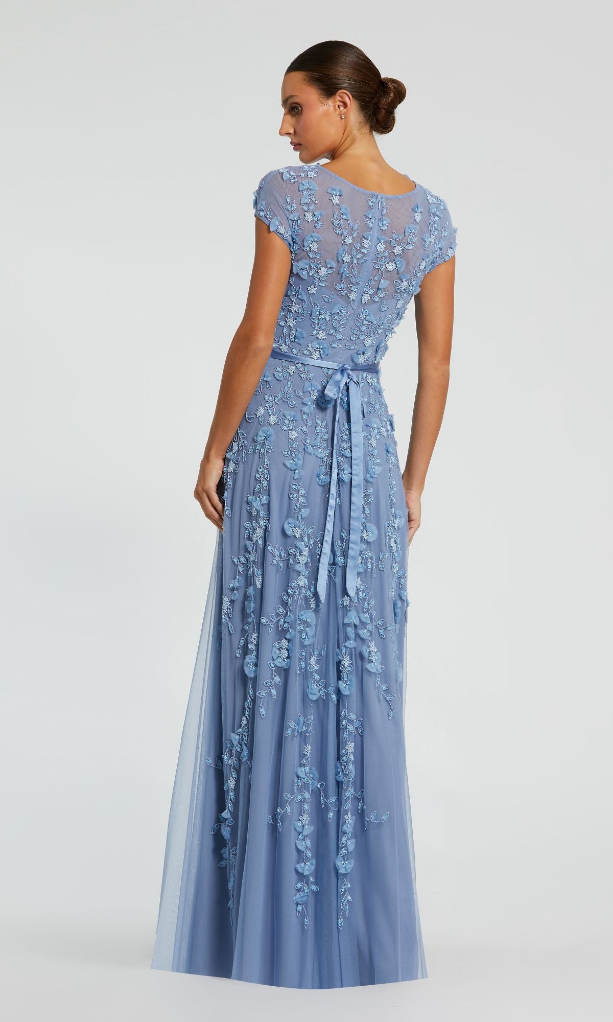 Woman stands with her back to the camera in the Long Sky Blue Formal Dress: Mac Duggal 14521, featuring floral embroidery, a waist sash, and short sleeves for an elegant look.