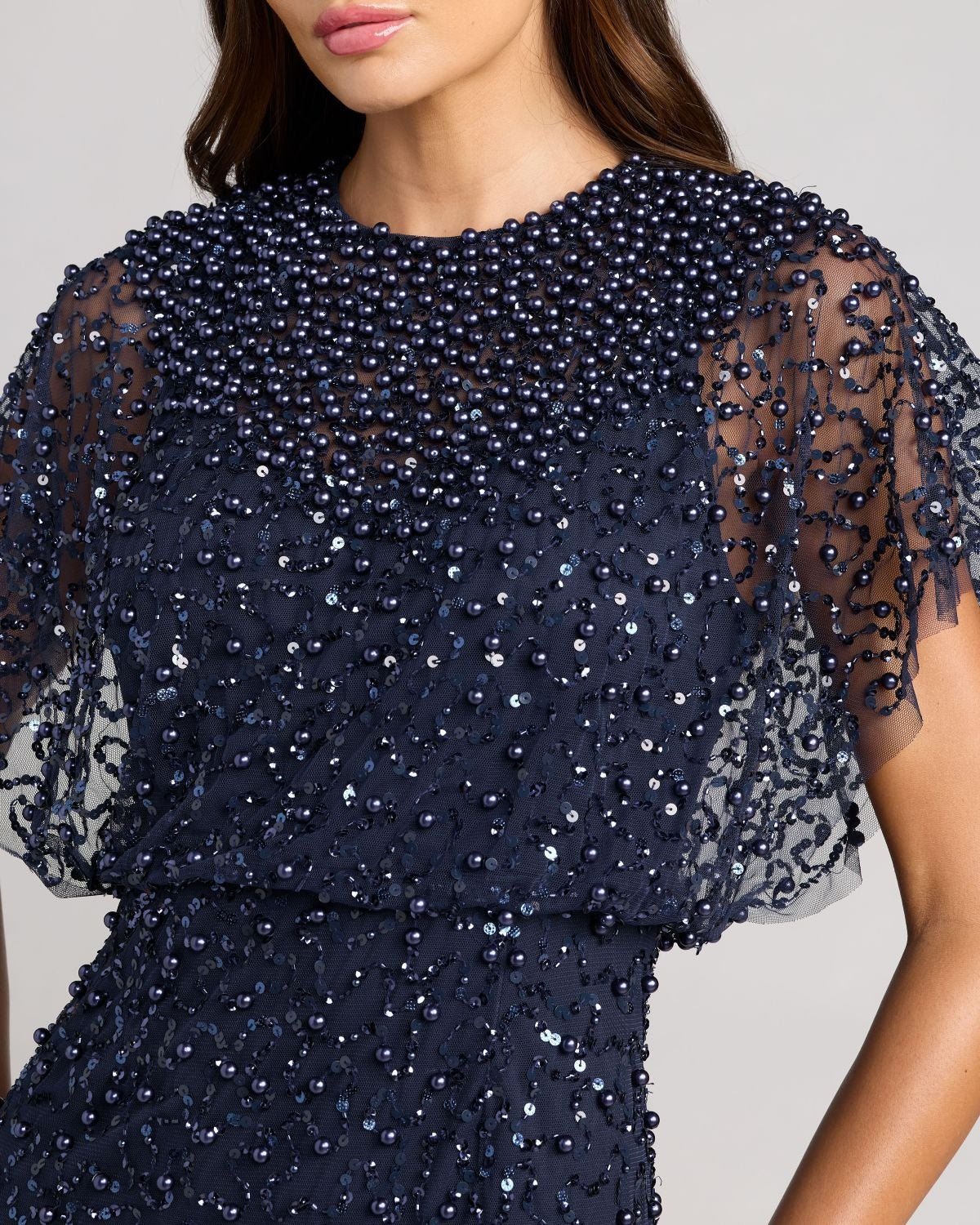 A woman wears the Mac Duggal 14523 Beaded Long Nude Evening Gown featuring sheer sleeves, a sheer bodice, and pearl and sequin embellishments.