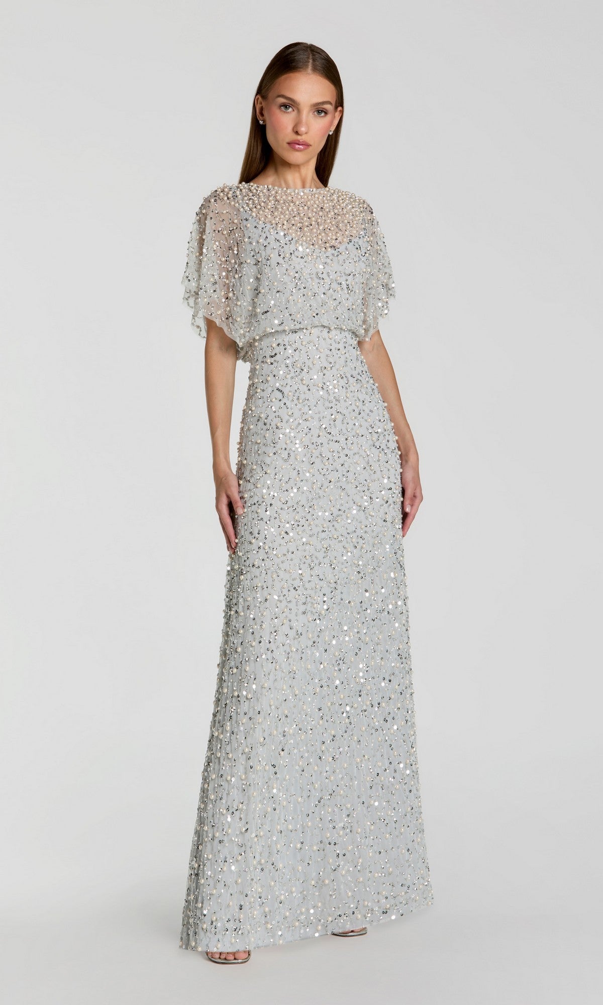 A woman wears the Beaded Long Nude Evening Gown: Mac Duggal 14523, a floor-length dress with silver sequins, a sheer embellished overlay, and short sleeves, standing against a plain light background.