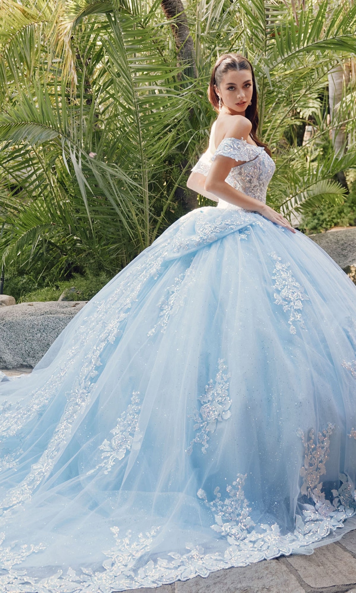 A woman stands outdoors among lush green plants, wearing the Quinceanera Dress By Juliet JT1458J—an off-the-shoulder, light blue tulle a-line gown with floral lace details.