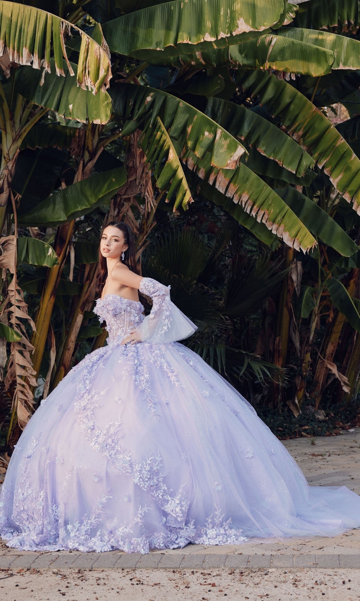 A woman in the Quinceanera Dress JT1459J by Juliet, an off-the-shoulder gown with floral details, stands on a path in front of large green banana leaves.